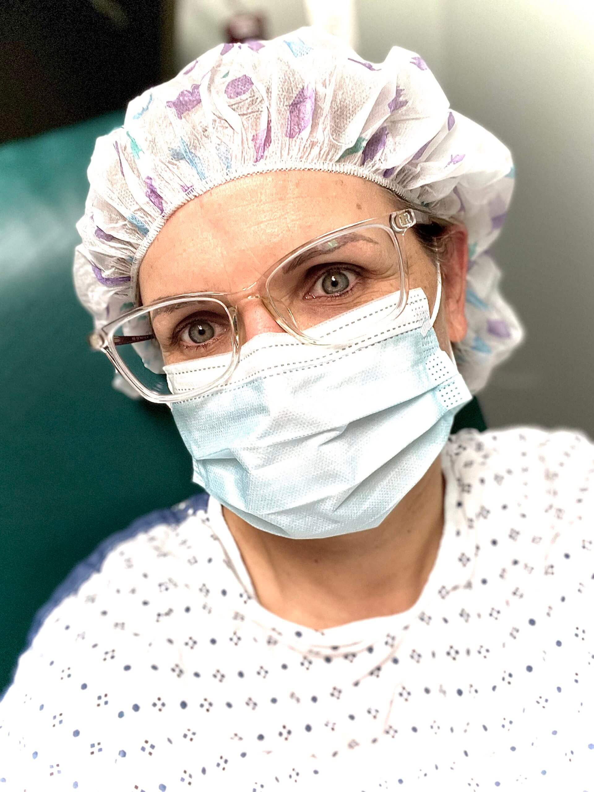 Color headshot of a white woman looking into camera. She id dressed for surgery in a gown, hairnet, mask, and is wearing clear-framed glasses.