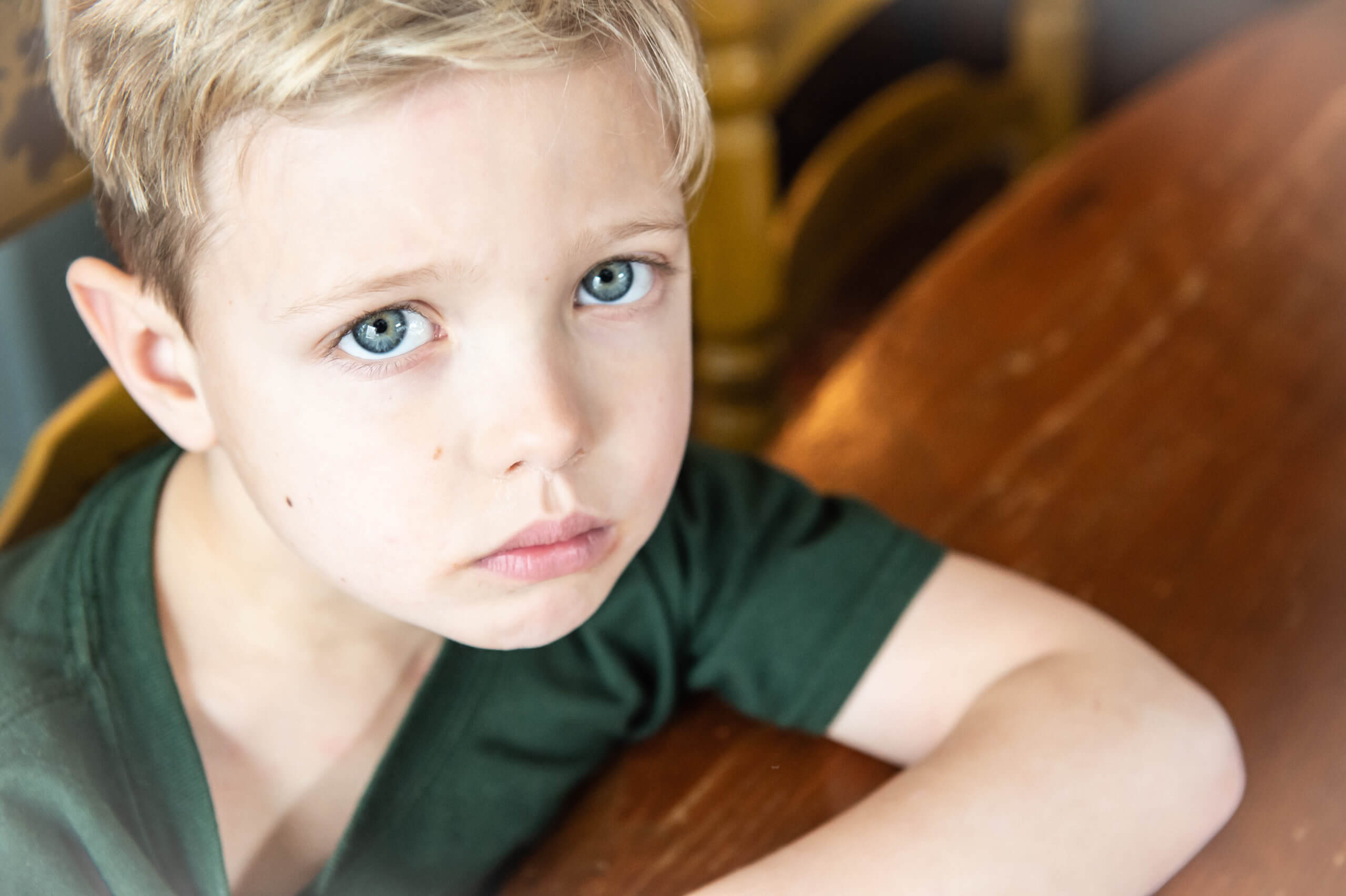 Color photo, close-up of 6 year old white boy with pale skin, blond hair, and green eyes (that match his green shirt) looking at the camera with a sad face.