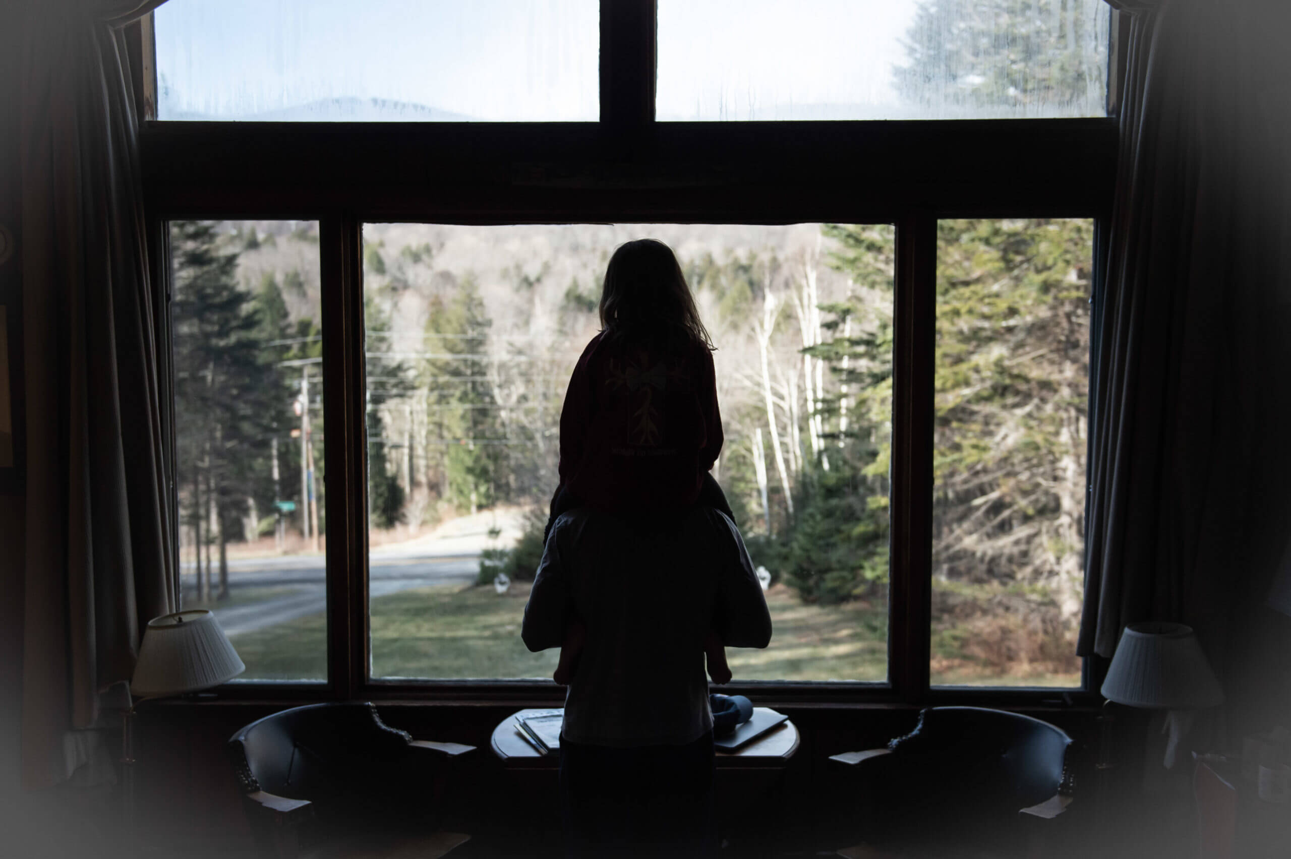 Silhouette of a girl on the shoulders of a man. They are looking out at a mountain and a late fall Vermont landscape through a large picture window.