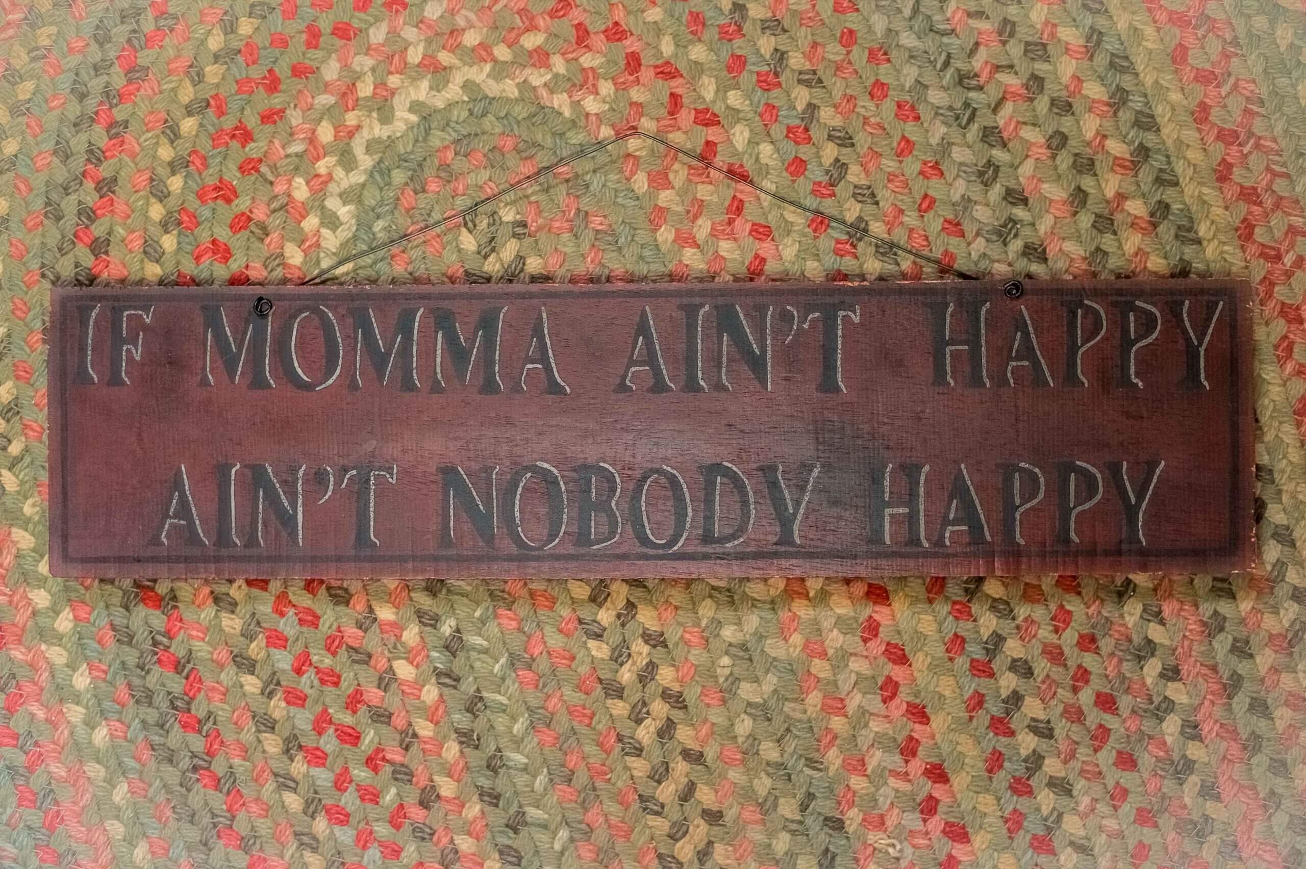 Color image of a wooden sign that reads "If Mamma Ain't Happy, Ain't Nobody Happy" on a laying on a braided rug.