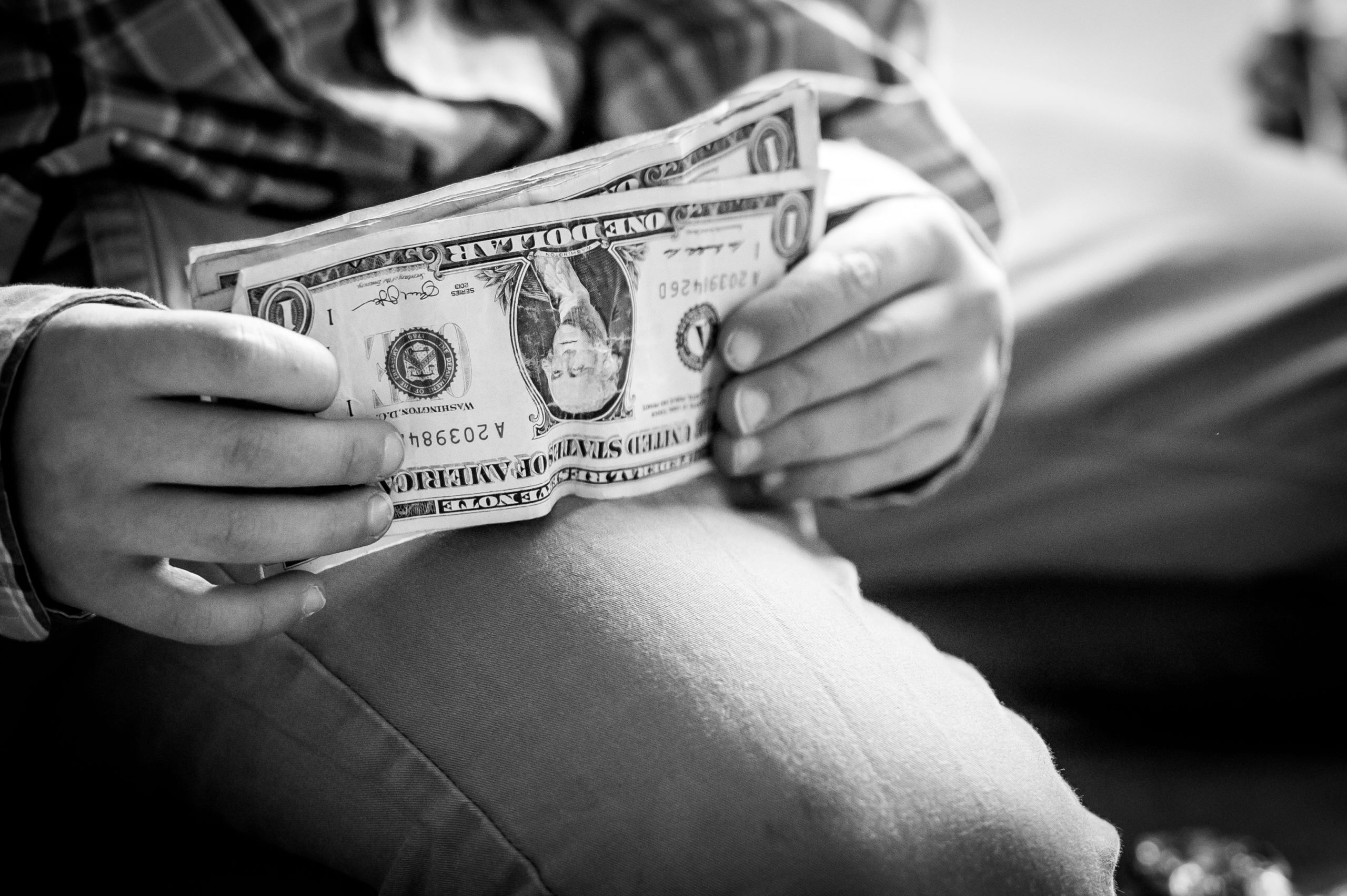 Black and white close-up photo of a little white boy holding up a stack of about ten one dollar bills. He's kneeling and has them resting on his thigh as if he's trying to straighten the stack.