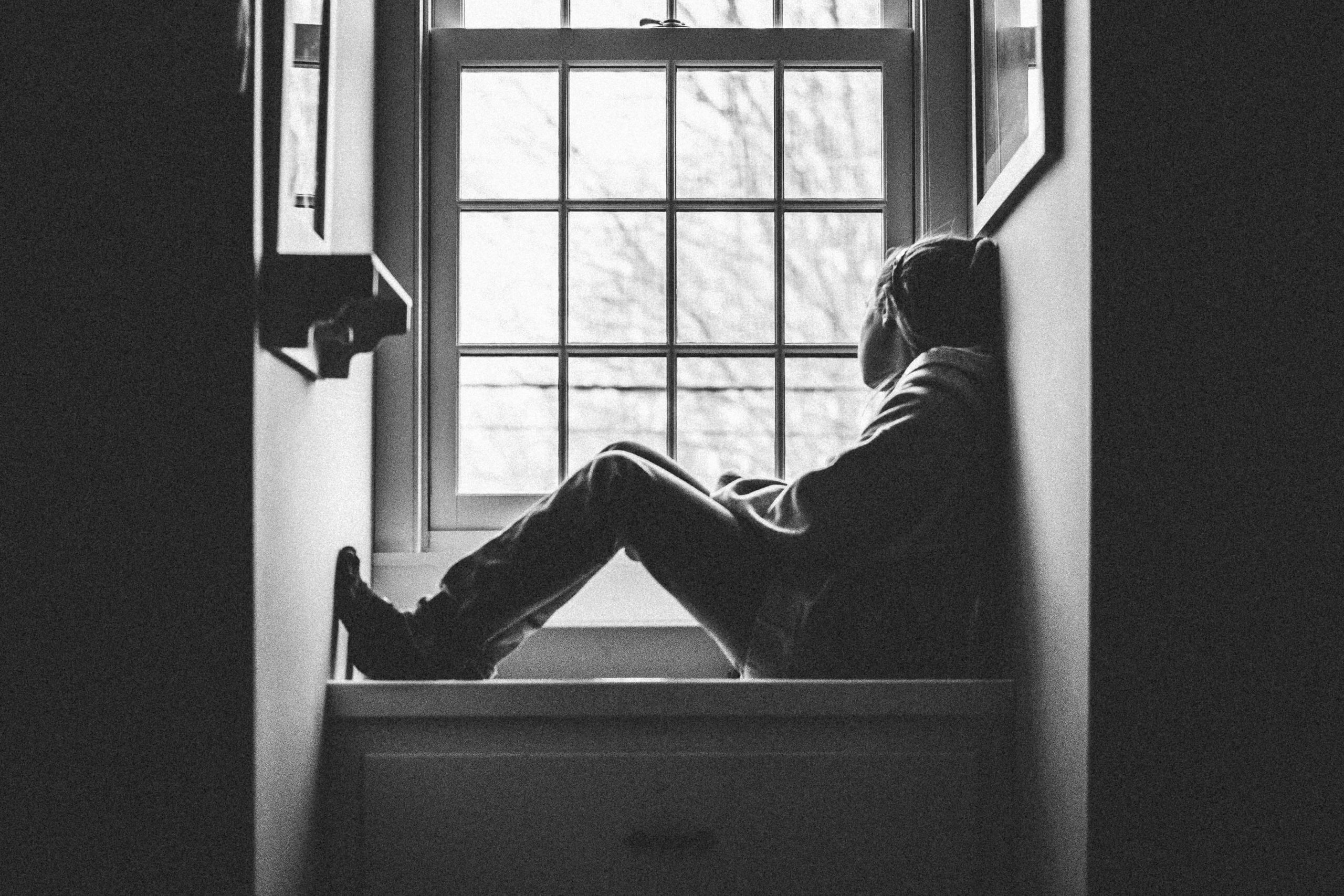 Black and white image of a white tween girl sitting on a window seat looking out the window.