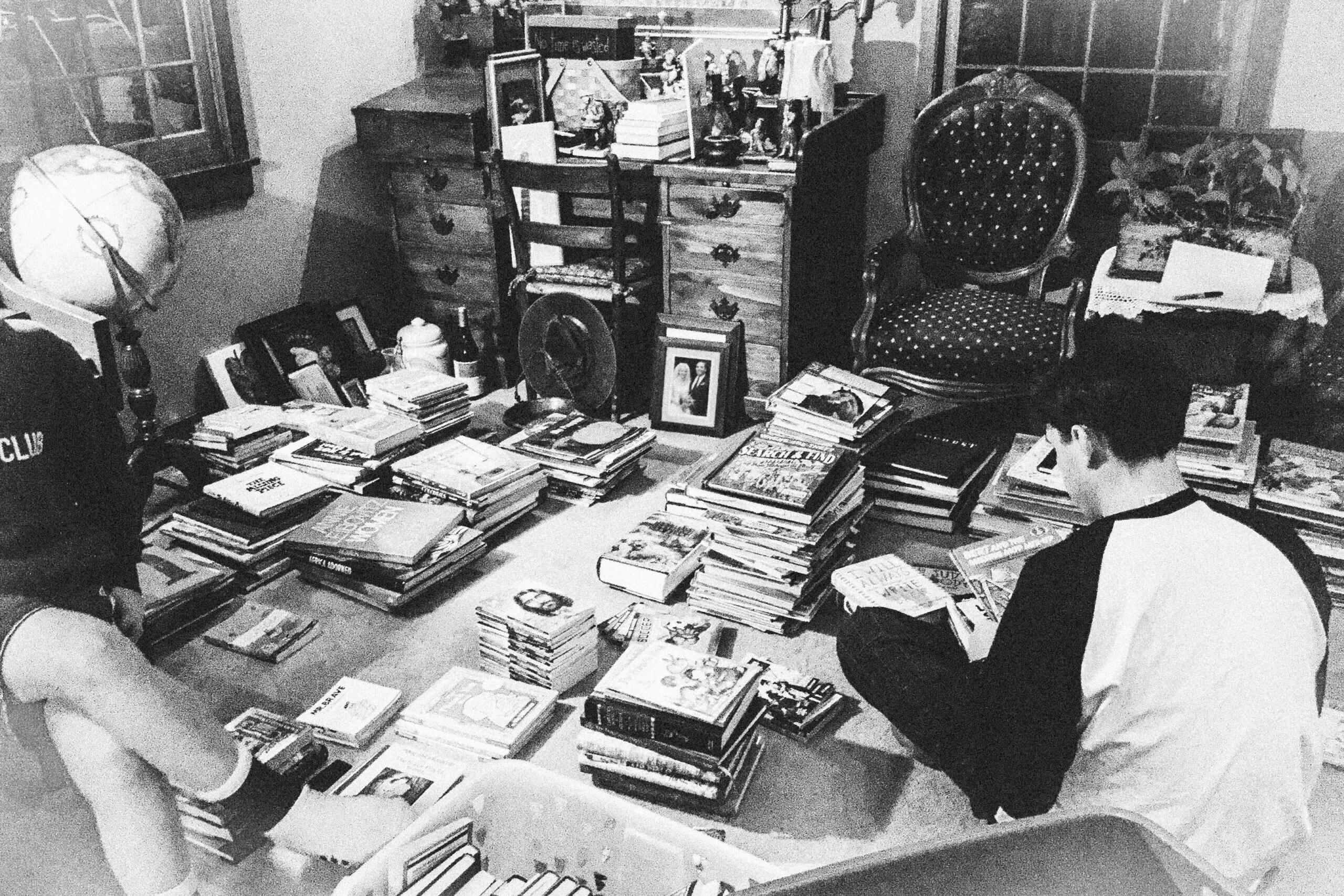 Black and white grainy image of a room filled with stacks of books and stuff. A white man sits on the floor and goes through the books.