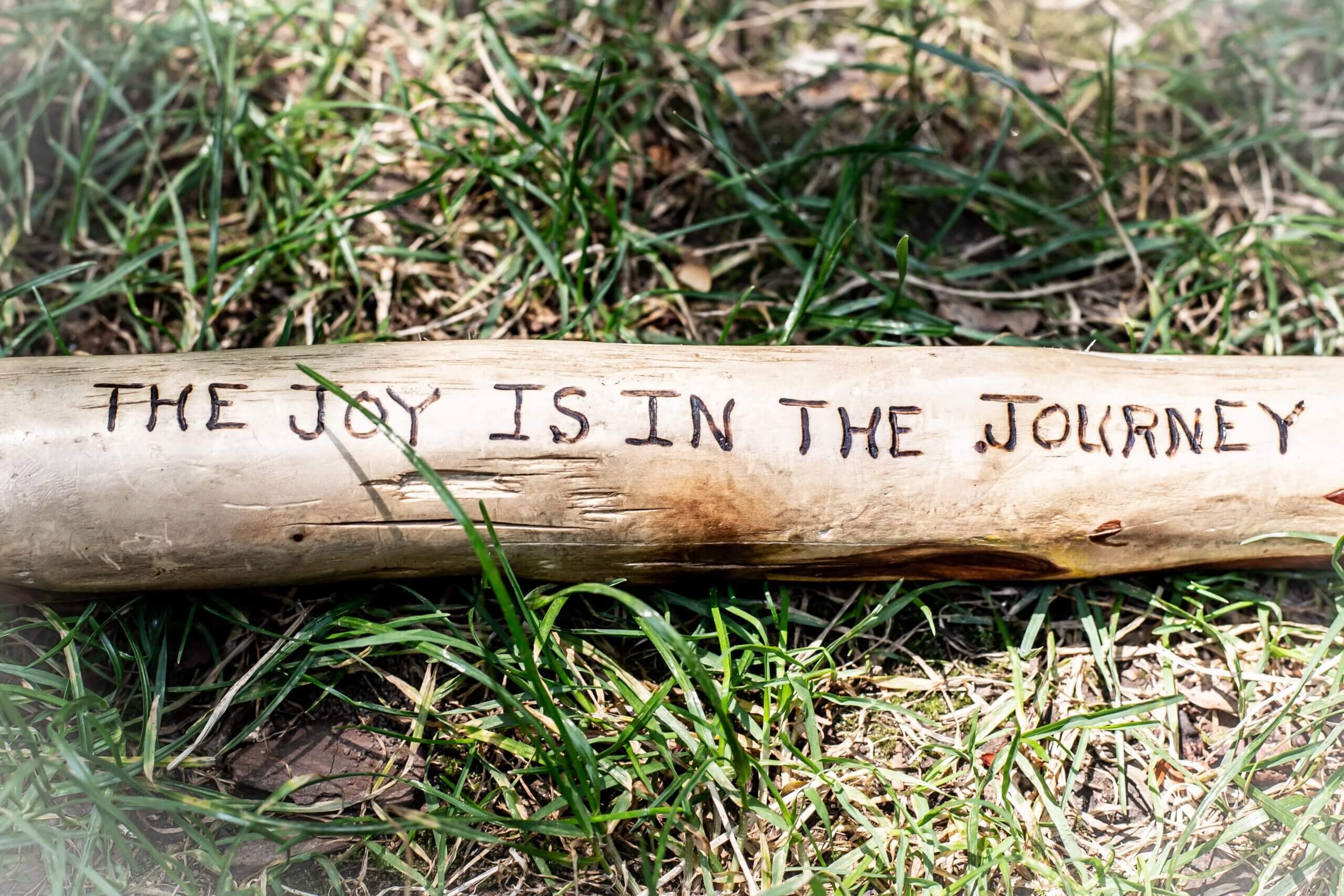 A piece of wood sitting on green grass that reads: THE JOY IS IN THE JOURNEY