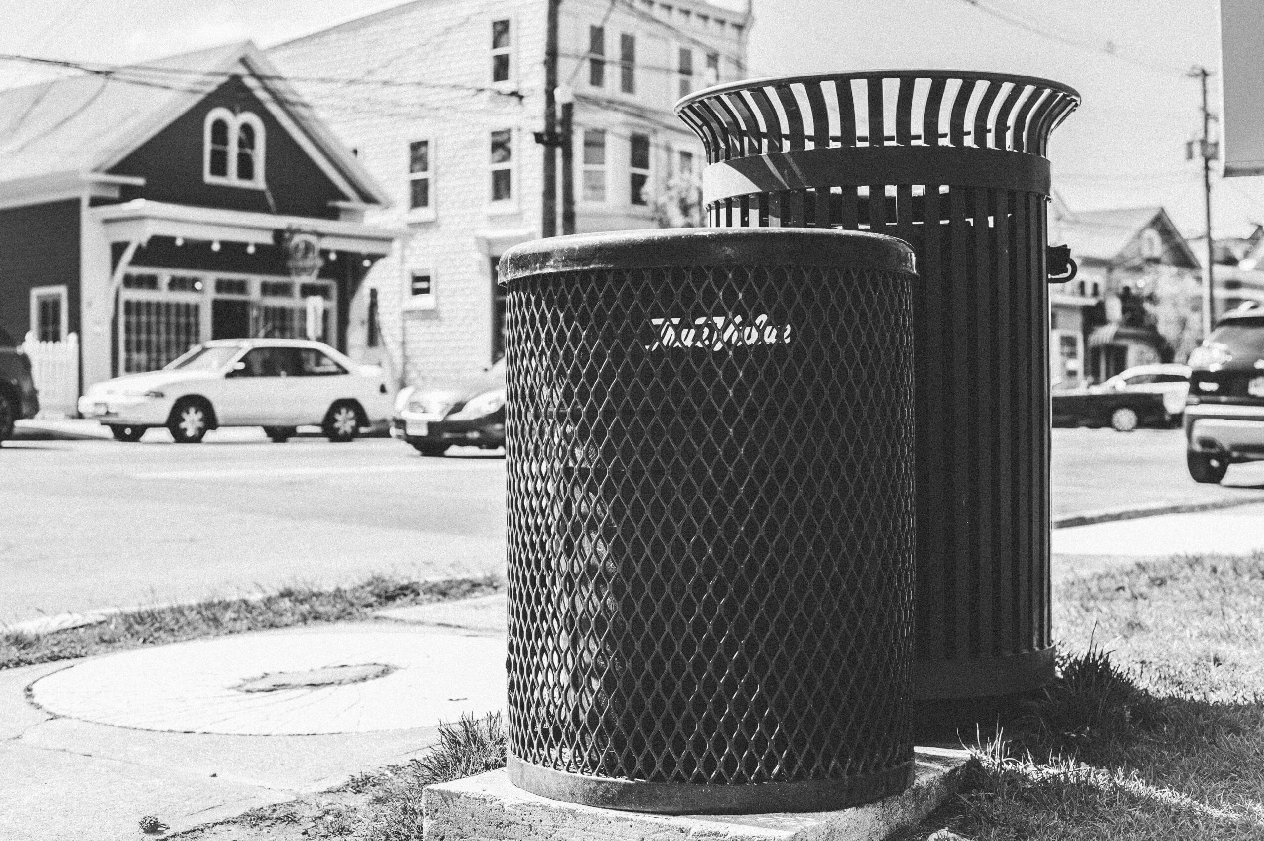 Black and white image of two metal circular trash cans on the corner of a sidewalk in a New England town.