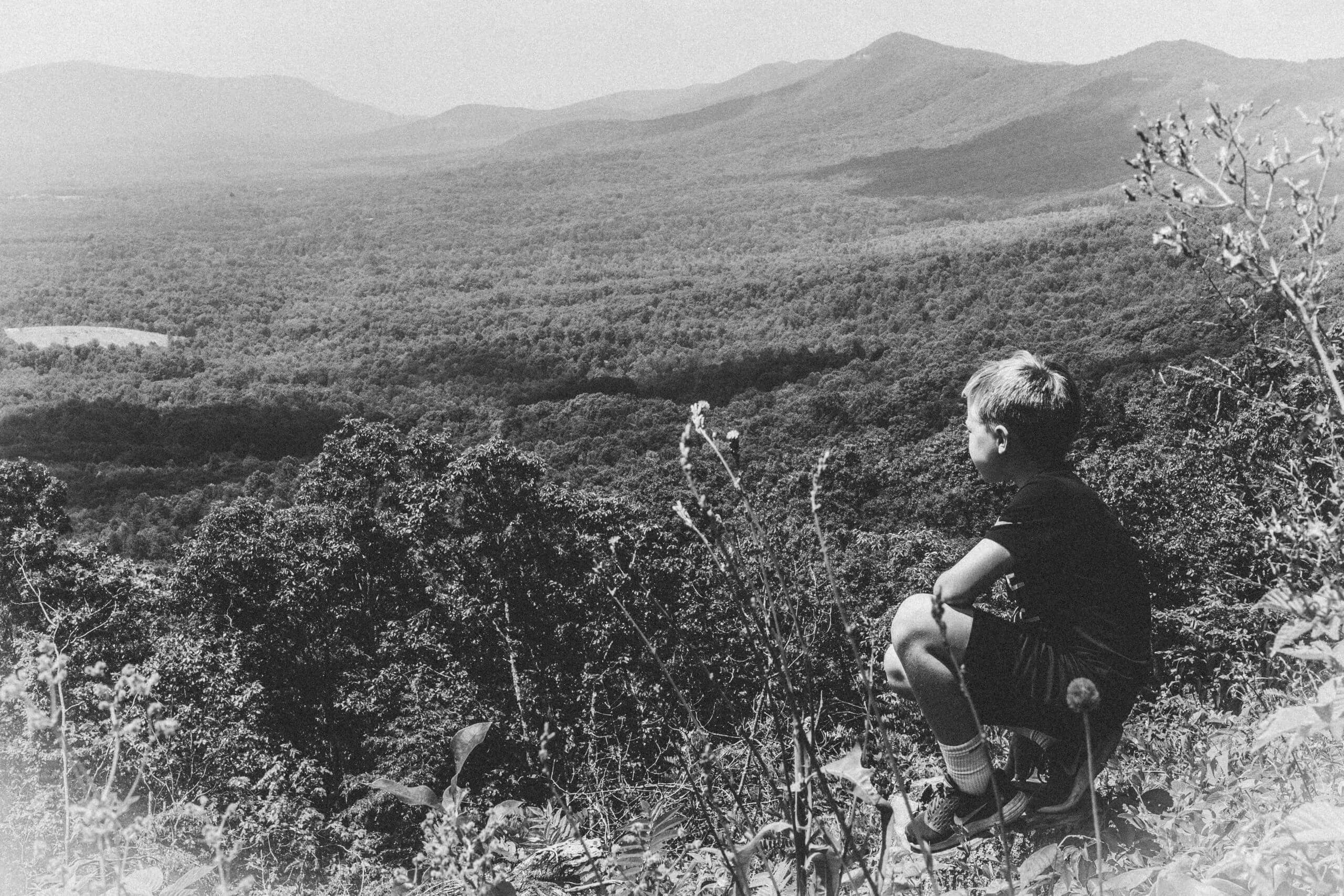 Graney black and white image of a grassy mountain scene with a white boy with blond hair crouched down in the bottom right third of the frame looking out at the landscape.