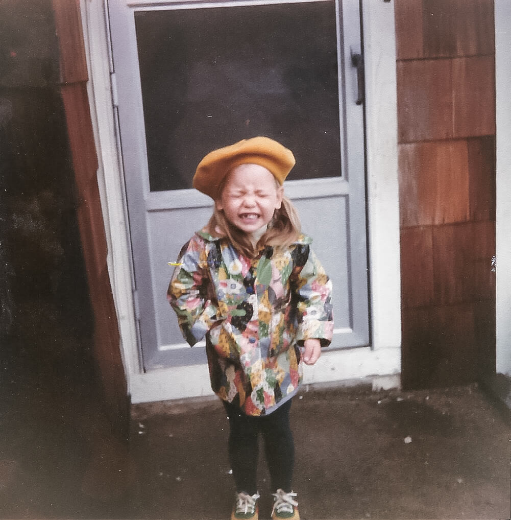 Old 1 by 1 color photo from the 80's of a little white five-year-old girl with a missing front tooth, smiling for the camera. She has on a raincoat that looks like patchwork and a yellow beret.