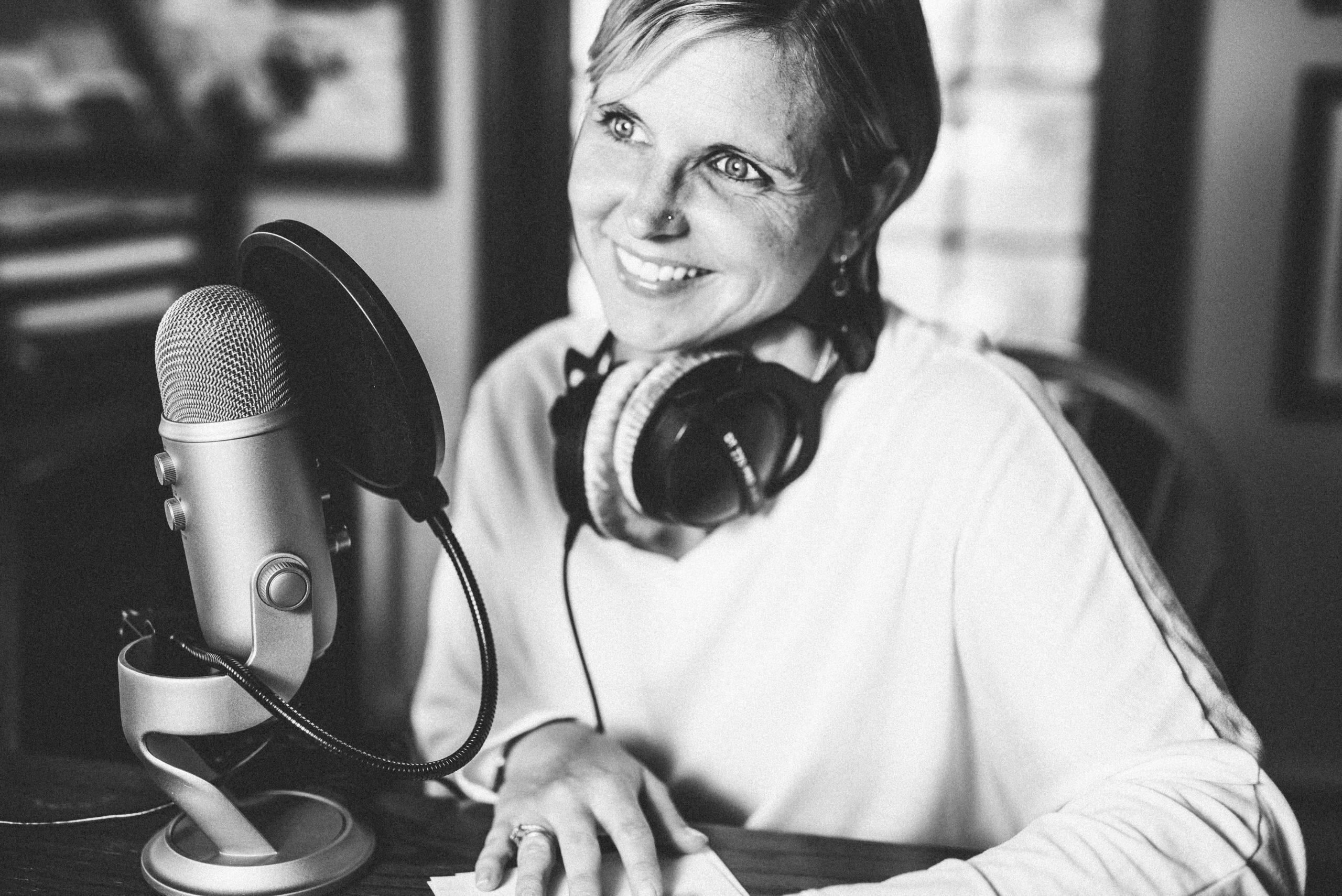 A black and white image of a white 40-something year old woman with short hair looking off into the distance. She has recording studio headphones around her neck and is sitting in front of a microphone with a pop filter.