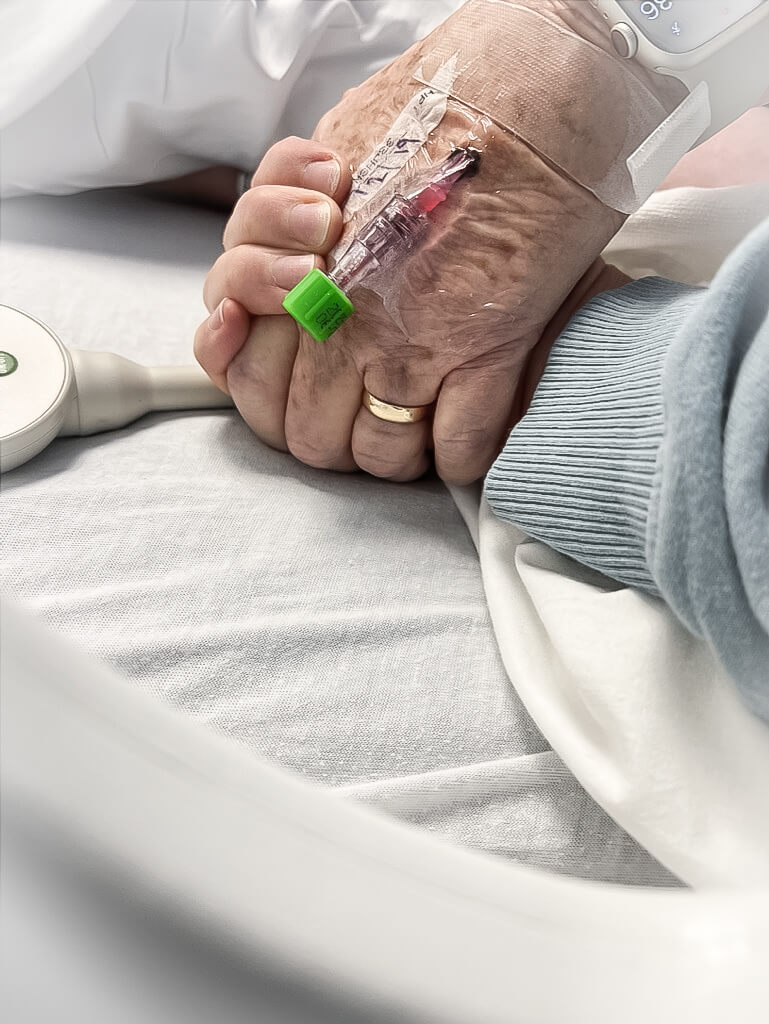 Close up color image of a younger woman holding an older woman's hand in a hospital bed. The older woman has an IV in the back of her hand.