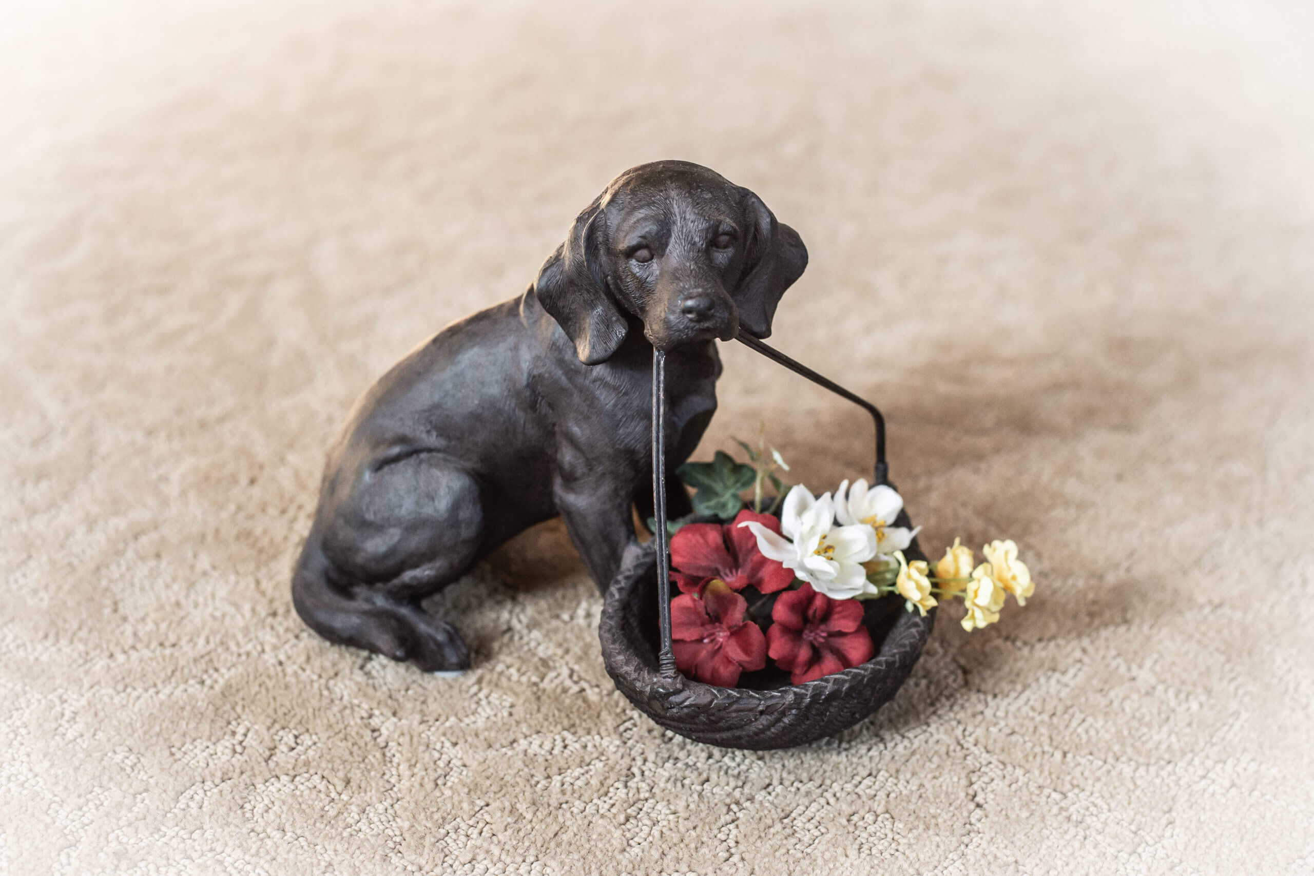 Color image of a dark brown dog figurine with a bucket in its mouth. The dog is in a seated position and the basket has small fake red, yellow, and white flowers in it.