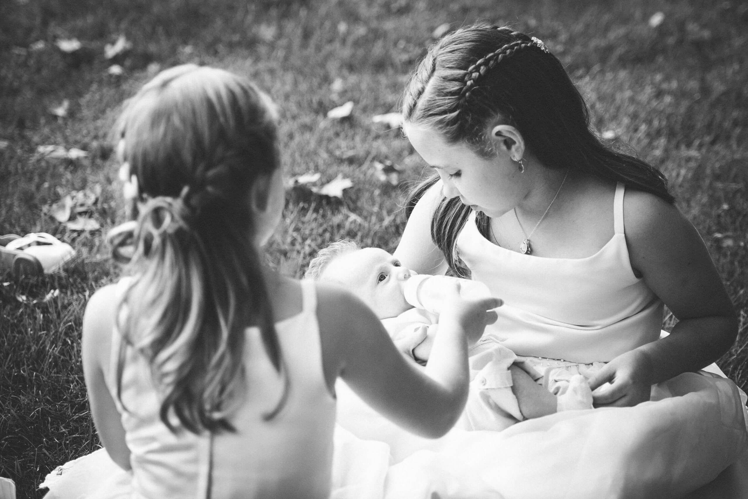 Black and white image of two young girls in pretty dresses. One little white girl is holding a baby in her lap, another little white girl is holding the bottle for the little white baby in the lap of the other girl.