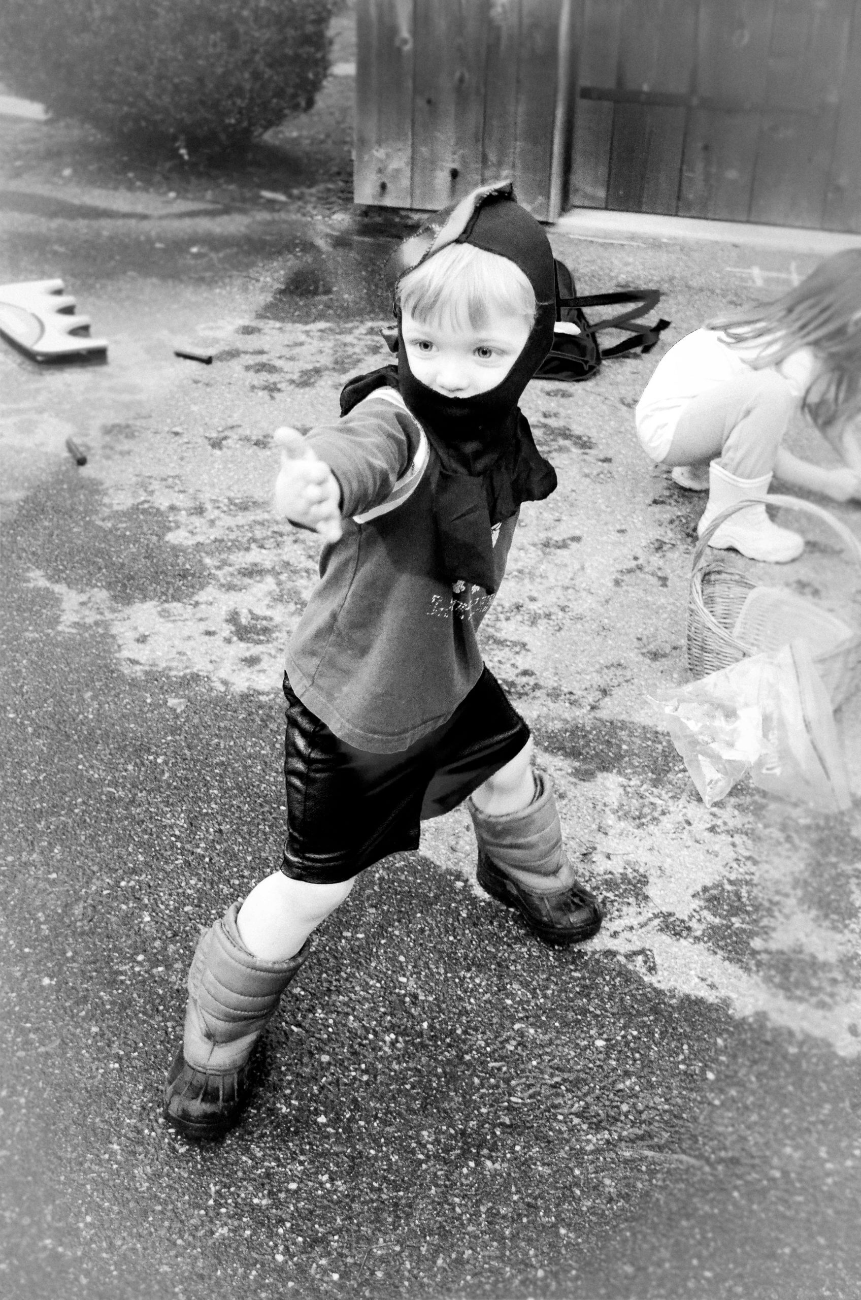 Black and white image of a white little boy dressed in a ninja hat doing a ninja move in shorts and snow boots.