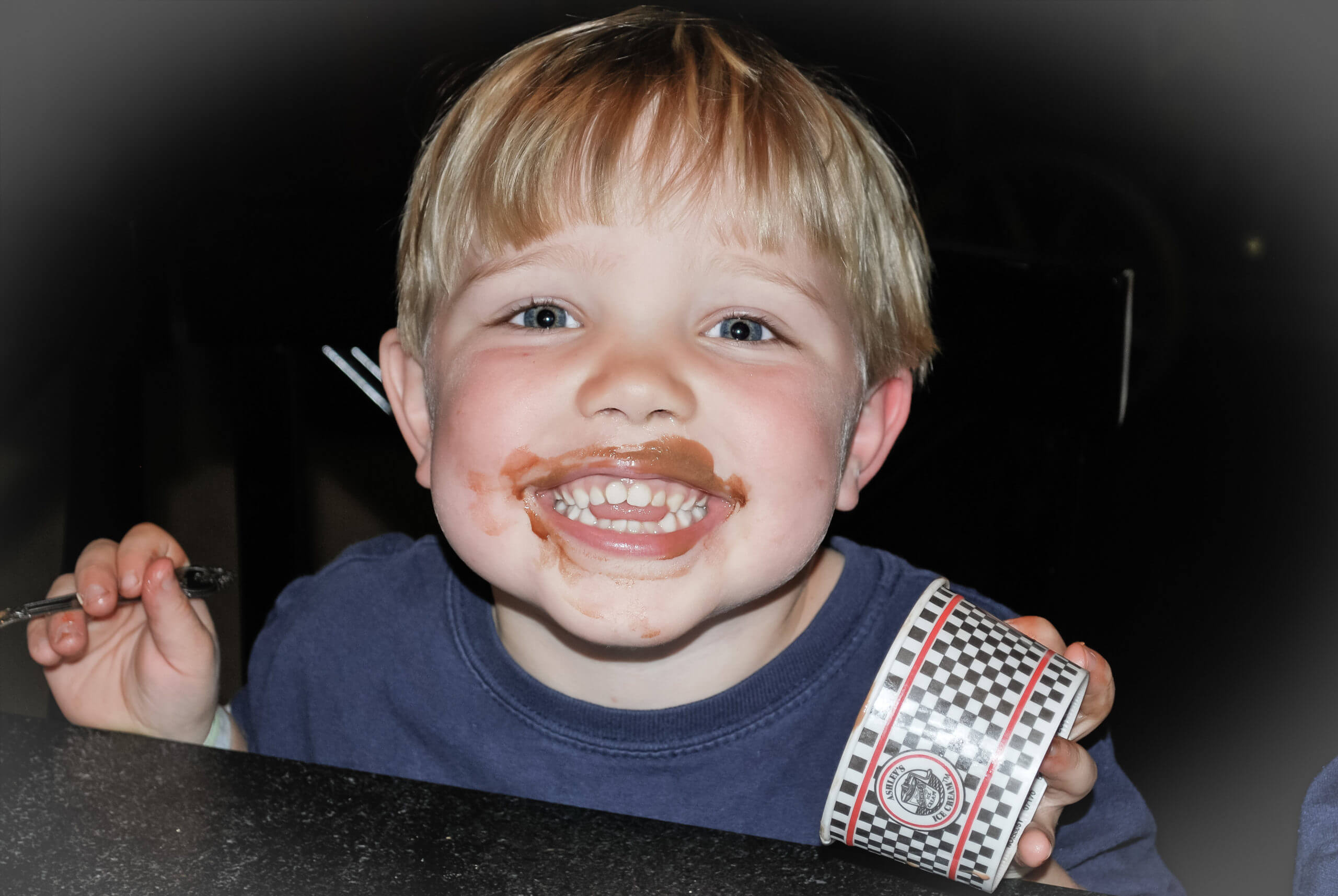 Close up image of a six-year-old white boy smiling with a mouth covered in chocolate ice cream. He's holding the empty checkered ice cream container in one hand, and his black plastic spoon in the other.