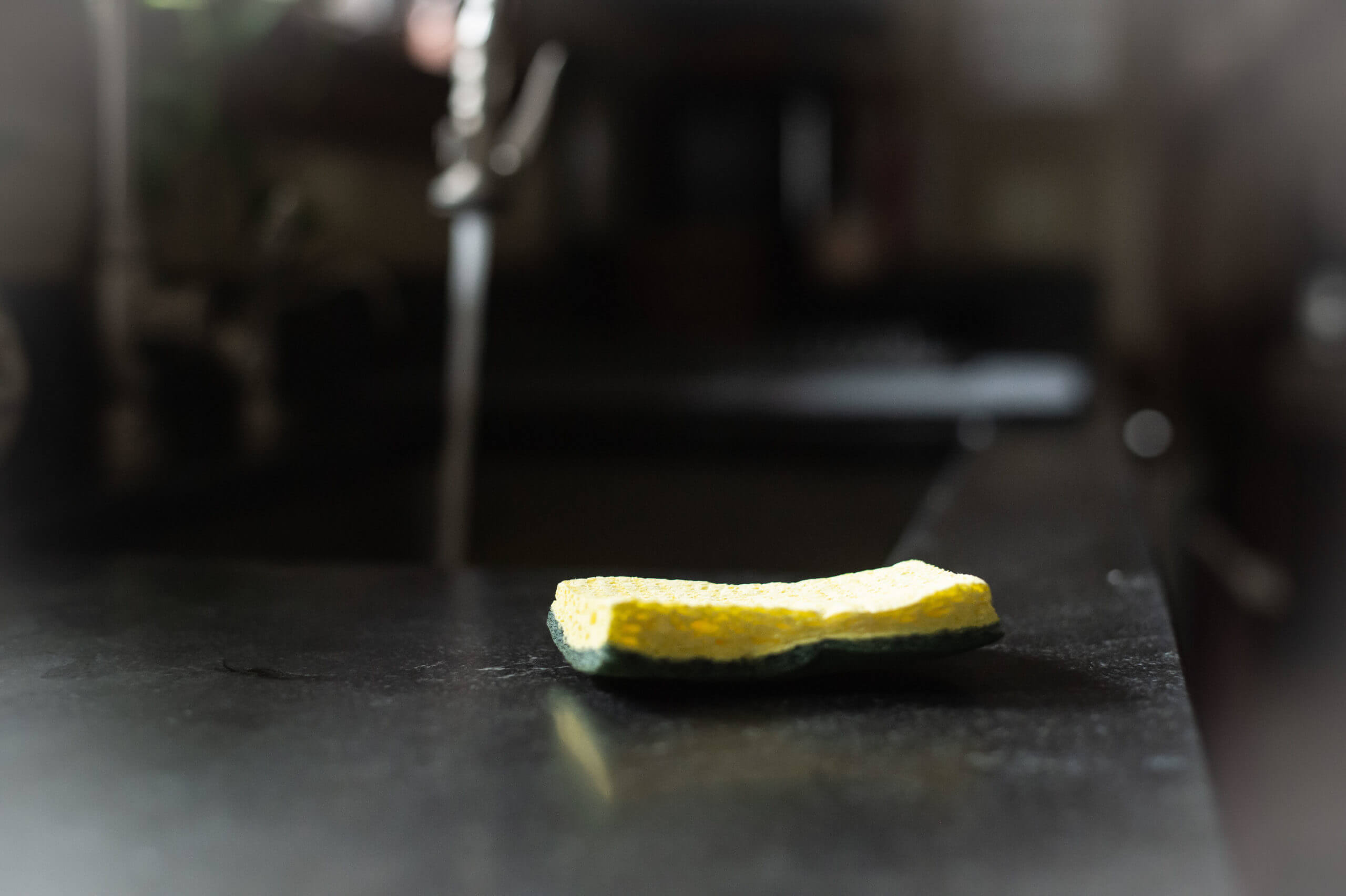Color image of a yellow sponge on a black countertop next to a kitchen sink. The faucet is running.