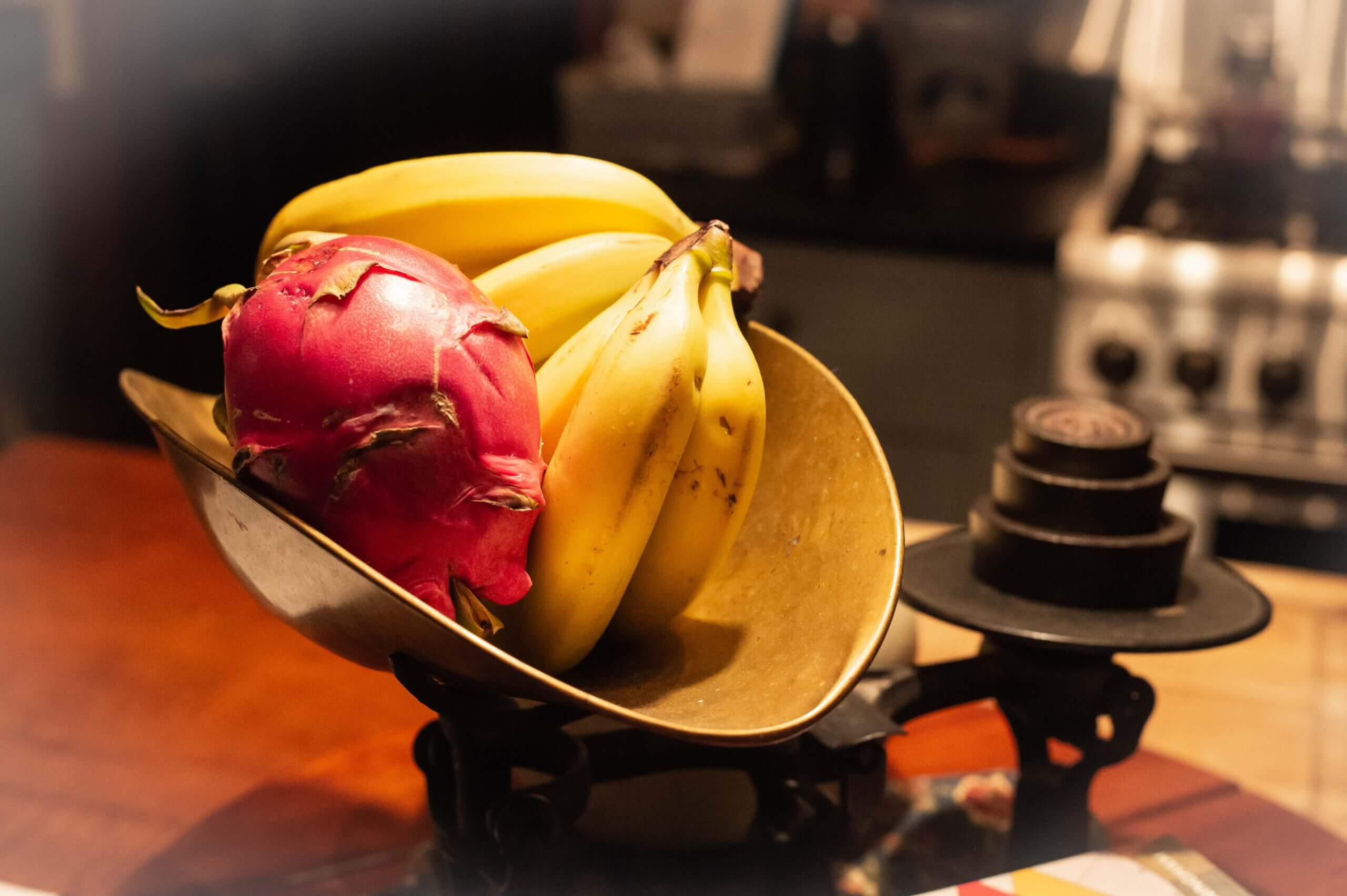Color image of an old fashioned scale in a kitchen, with yellow bananas and a red passion fruit on one side and circular weights on the other side.