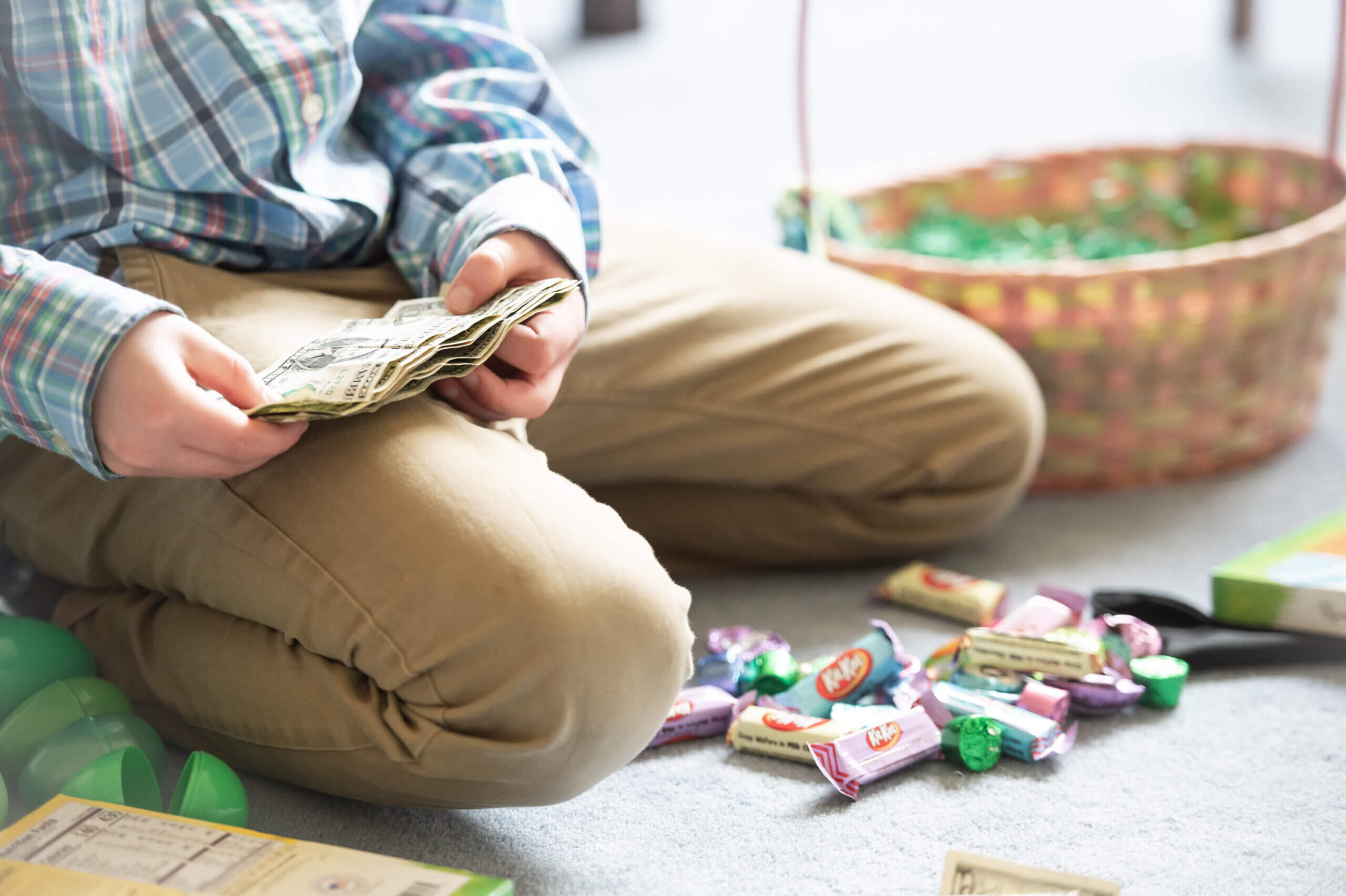 Color image of the bottom half of a young white boy in khakis and an Easter plaid shirt with a stack of small dollar bills in his hands and Easter candy and plastic Easter eggs around him on the light blue carpet.