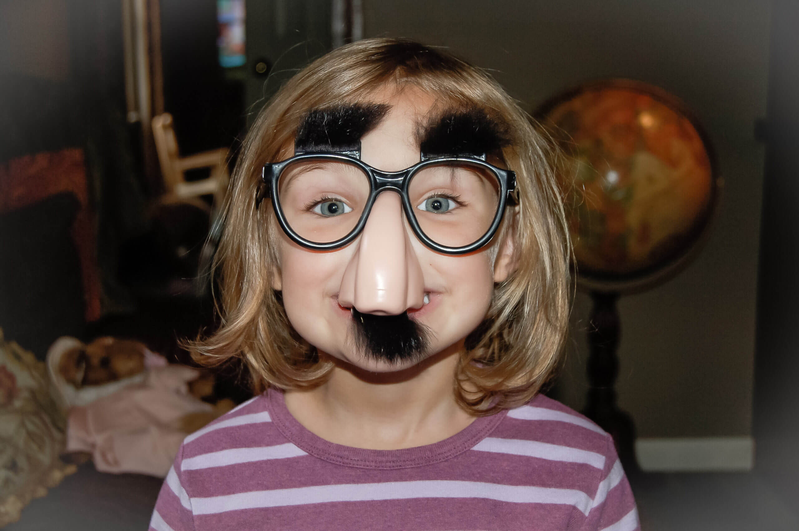 Color photo of a white little girl with a blond bob and Groucho Marx glasses on. The little girl has blue eyes and is smiling behind the big nose and fake moustache.