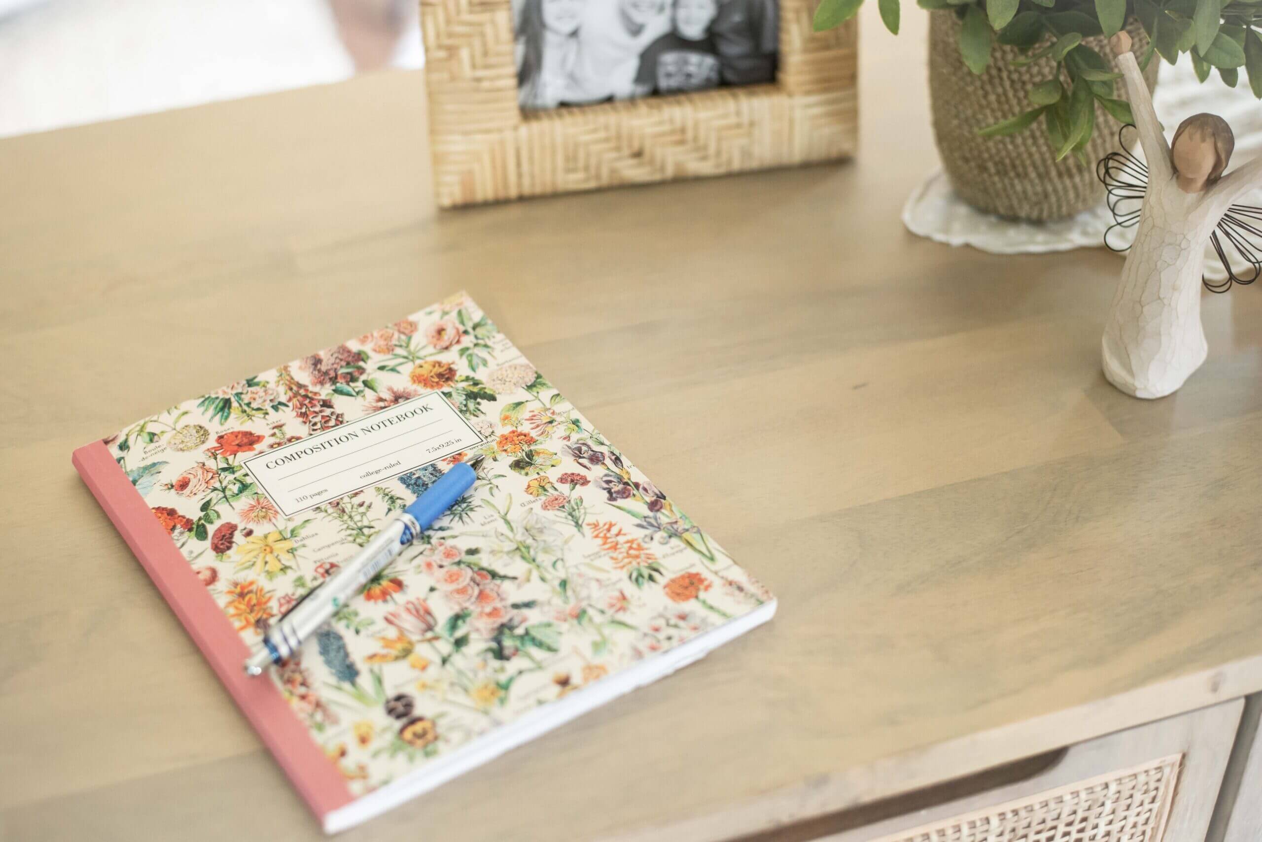 Color image of a pretty, flower-covered Composition Notebook with a blue pen on top of it, on a light wood desk. You can see a bit of a photo, a plant and a figurine in the background.