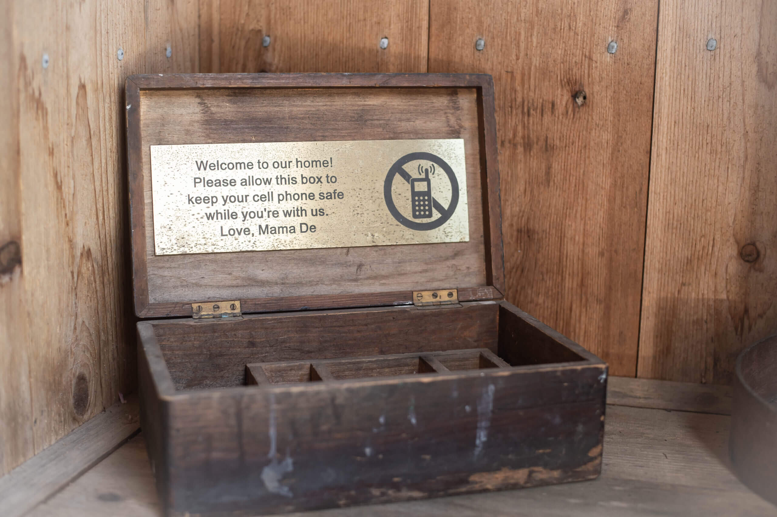 A color image of a wooden box opened up. There's a sign on the inside lid of the box that reads, "Welcome to our home! Please allow this box to keep your cell phone safe while you're with us. Love, Mama De." Next to the text there's a drawing of a cell phone inside of a circle with a cross going diagonally through it.