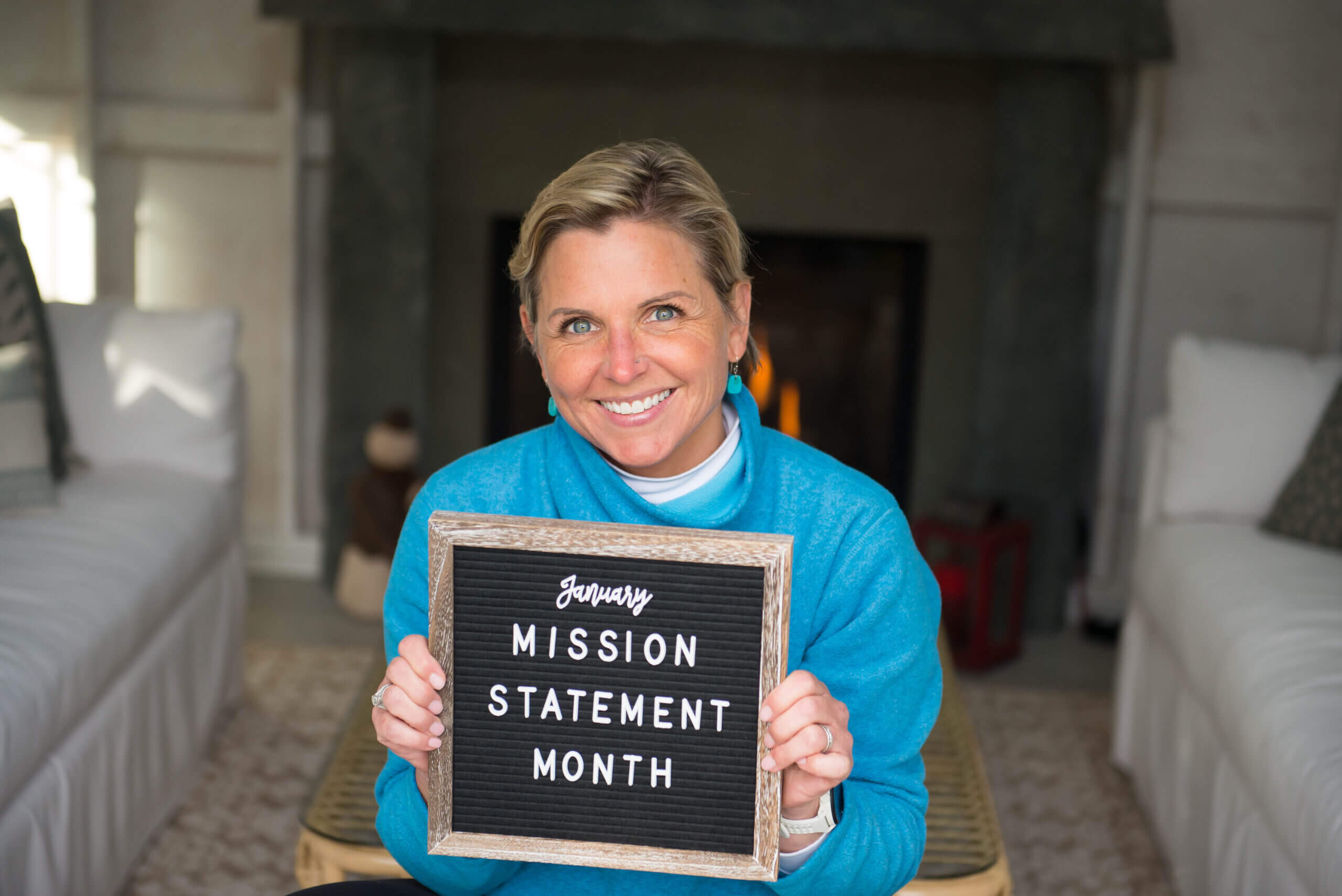 Mags smiling in front of a fireplace holding up a sign that says January Mission Statement Month