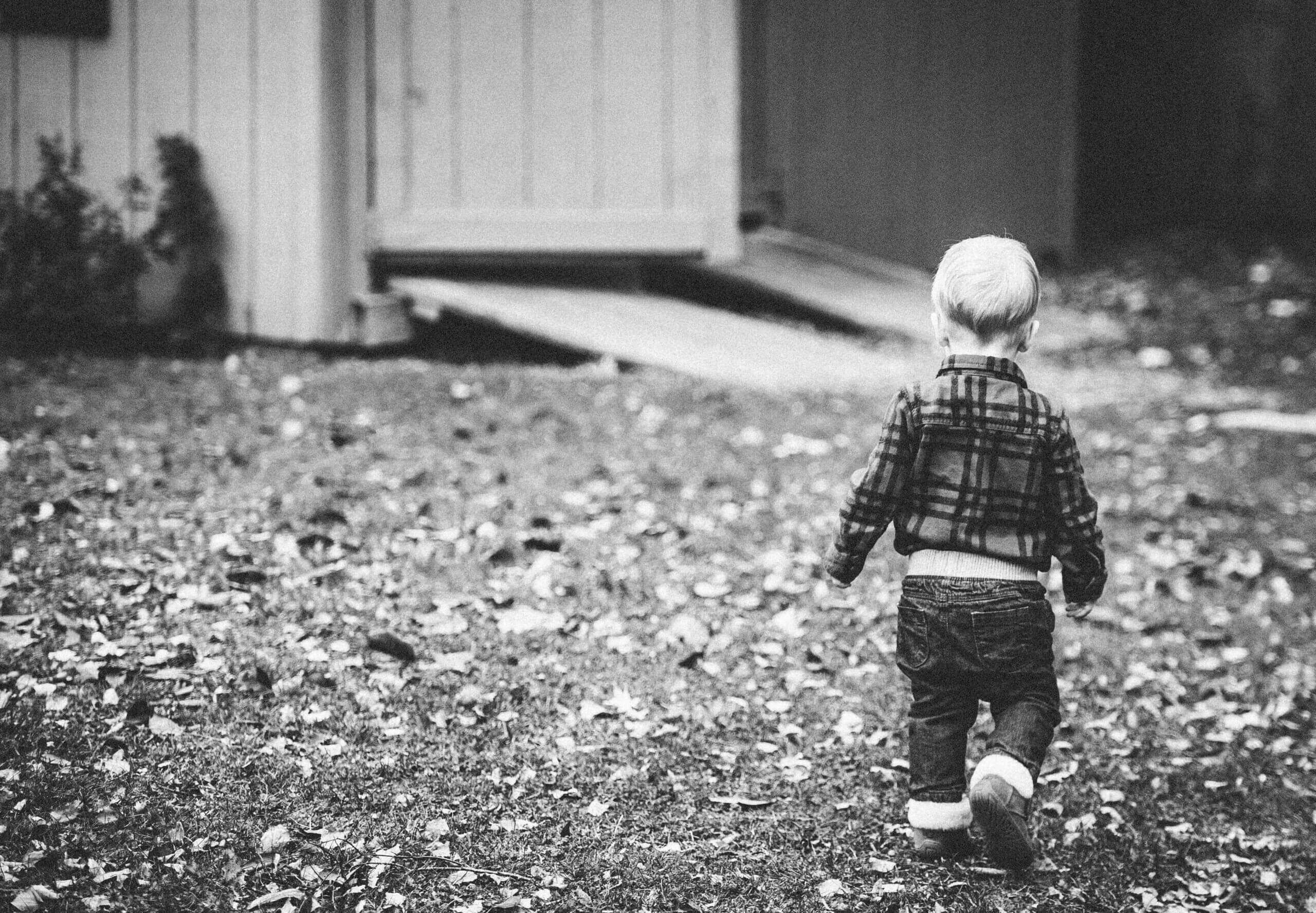 Black and white image of a white blond toddler walking away from the camera towards an open shed door.