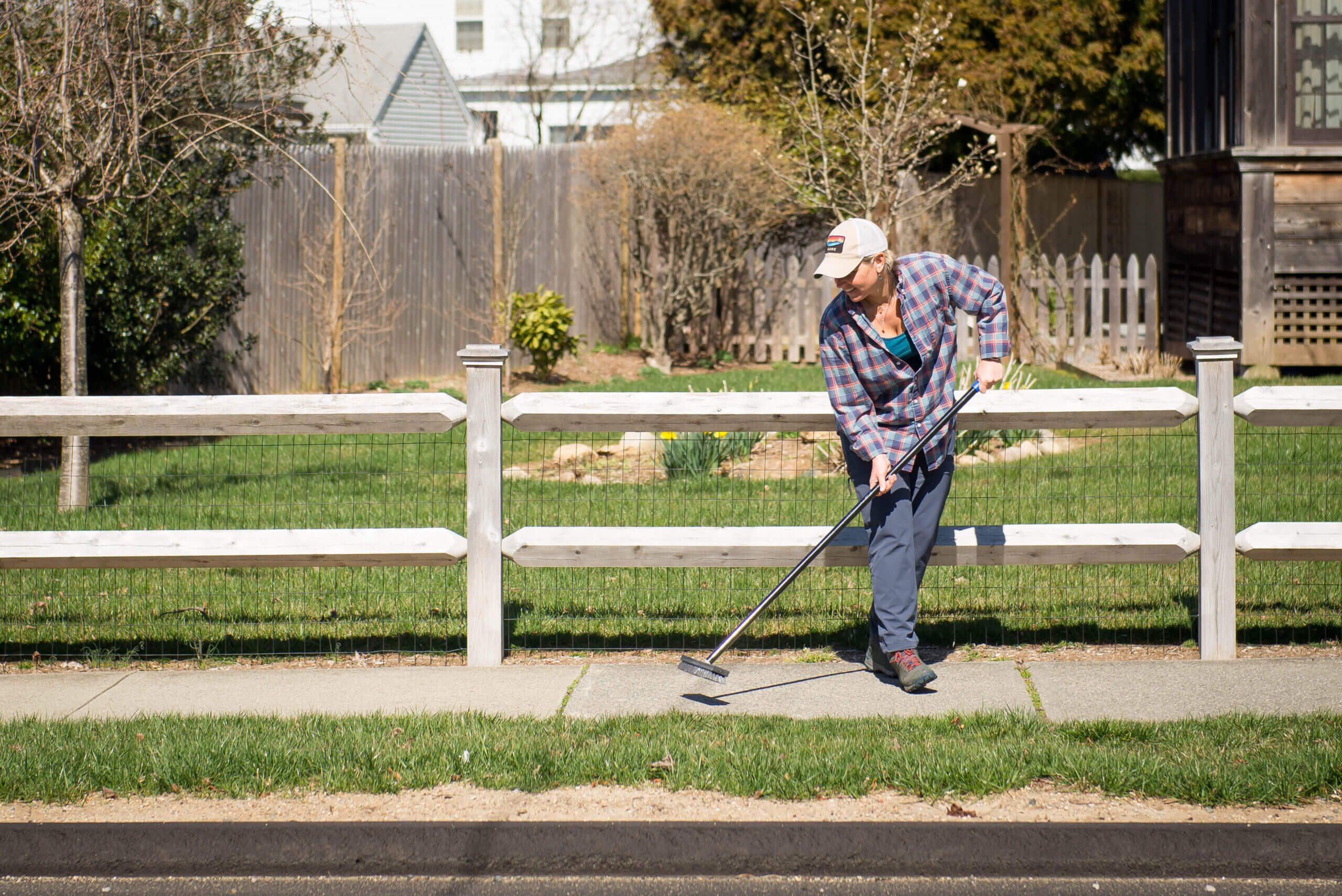 White woman sweeping a sidewalk in front of a fence.