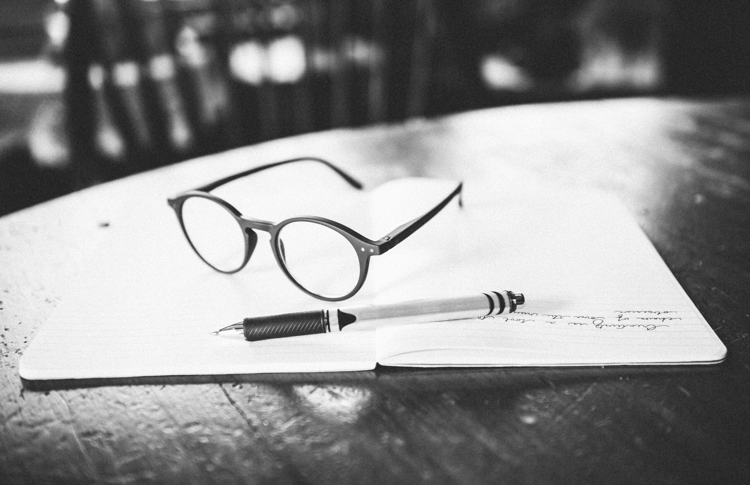 Black and white image of reading glasses and a pen lying on an open journal.