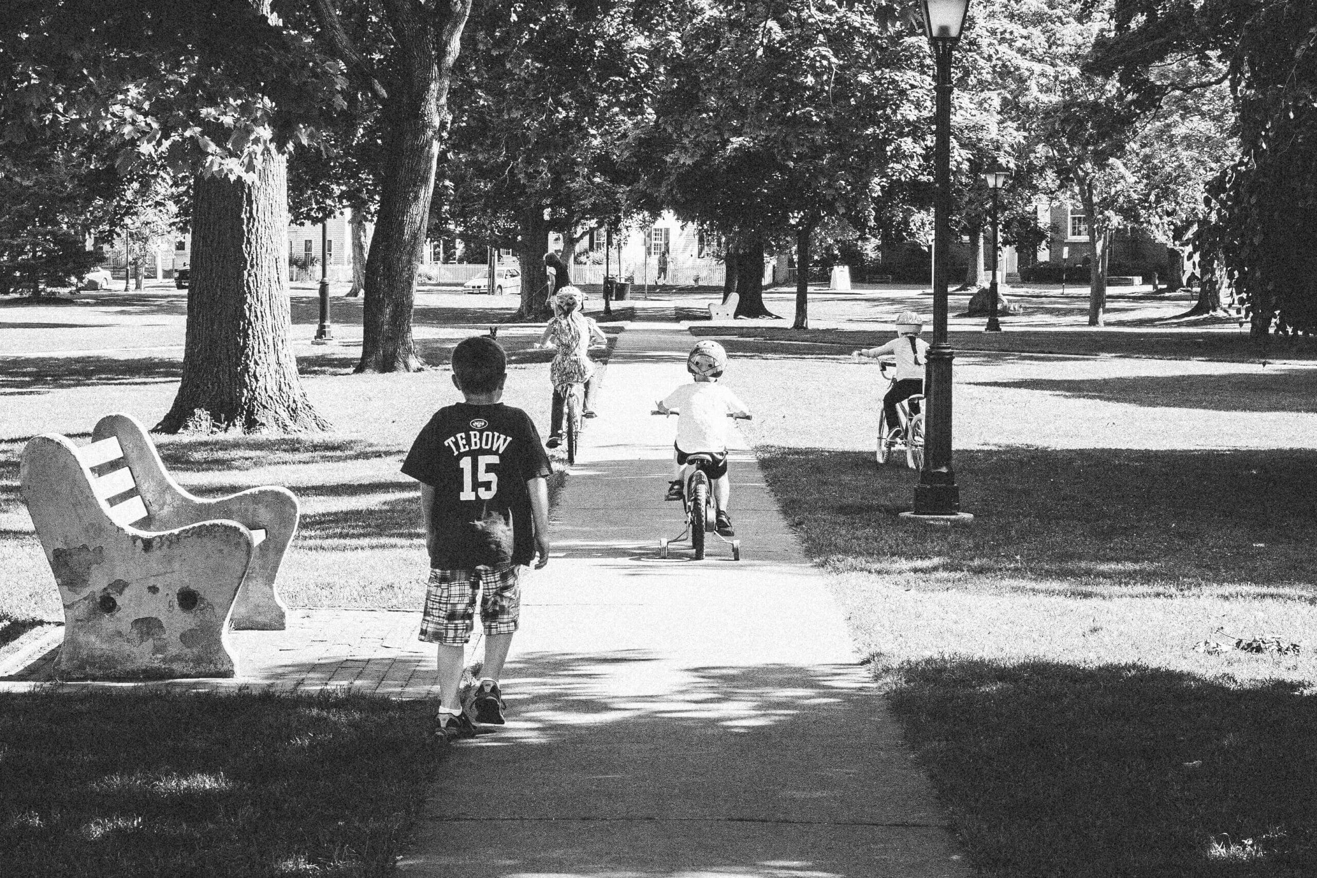 Black and white image of three kids riding bikes through a green and one kid walking. Their backs are to the camera.