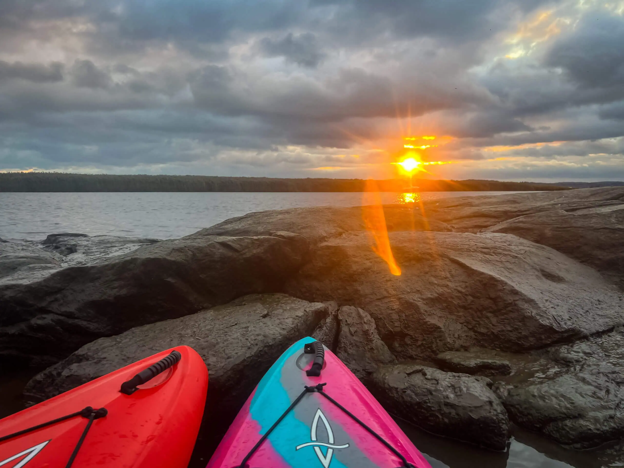 Color image of a sunset on the water with two kayaks in a jetty pointing at the sun. You can just see the tops of the boats.