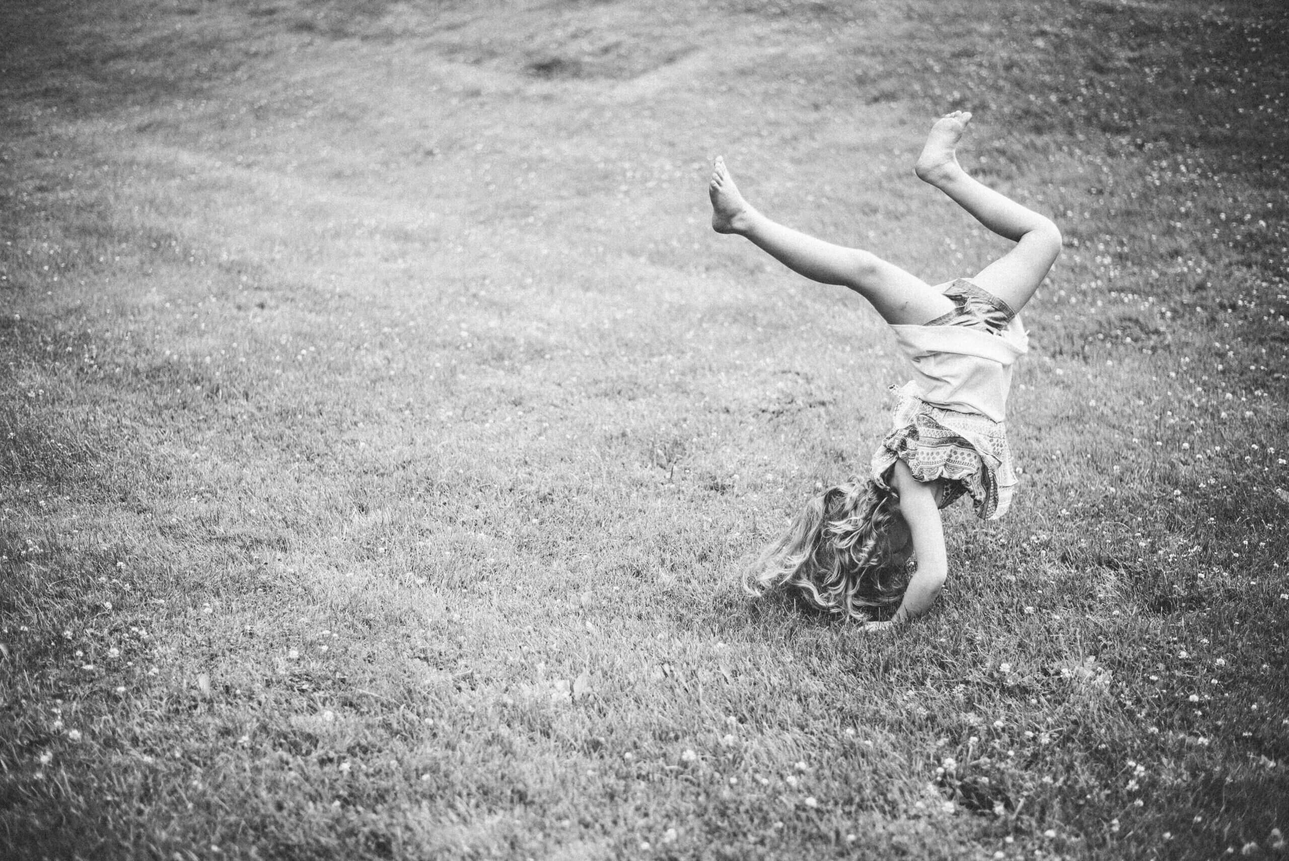 Black and white image of a little white girl upside down in the grass trying to land a cartwheel with her legs all crazy in the air.