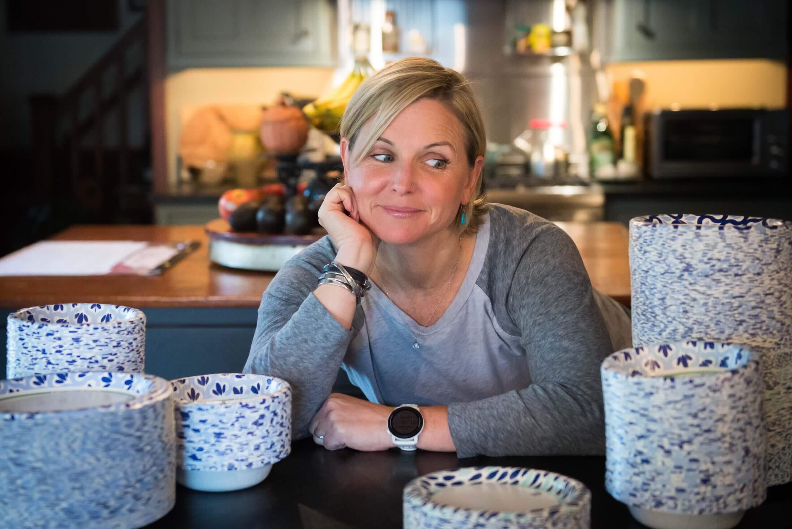 Color image of a white middle aged blond woman sitting amongst stacks of paper plates.