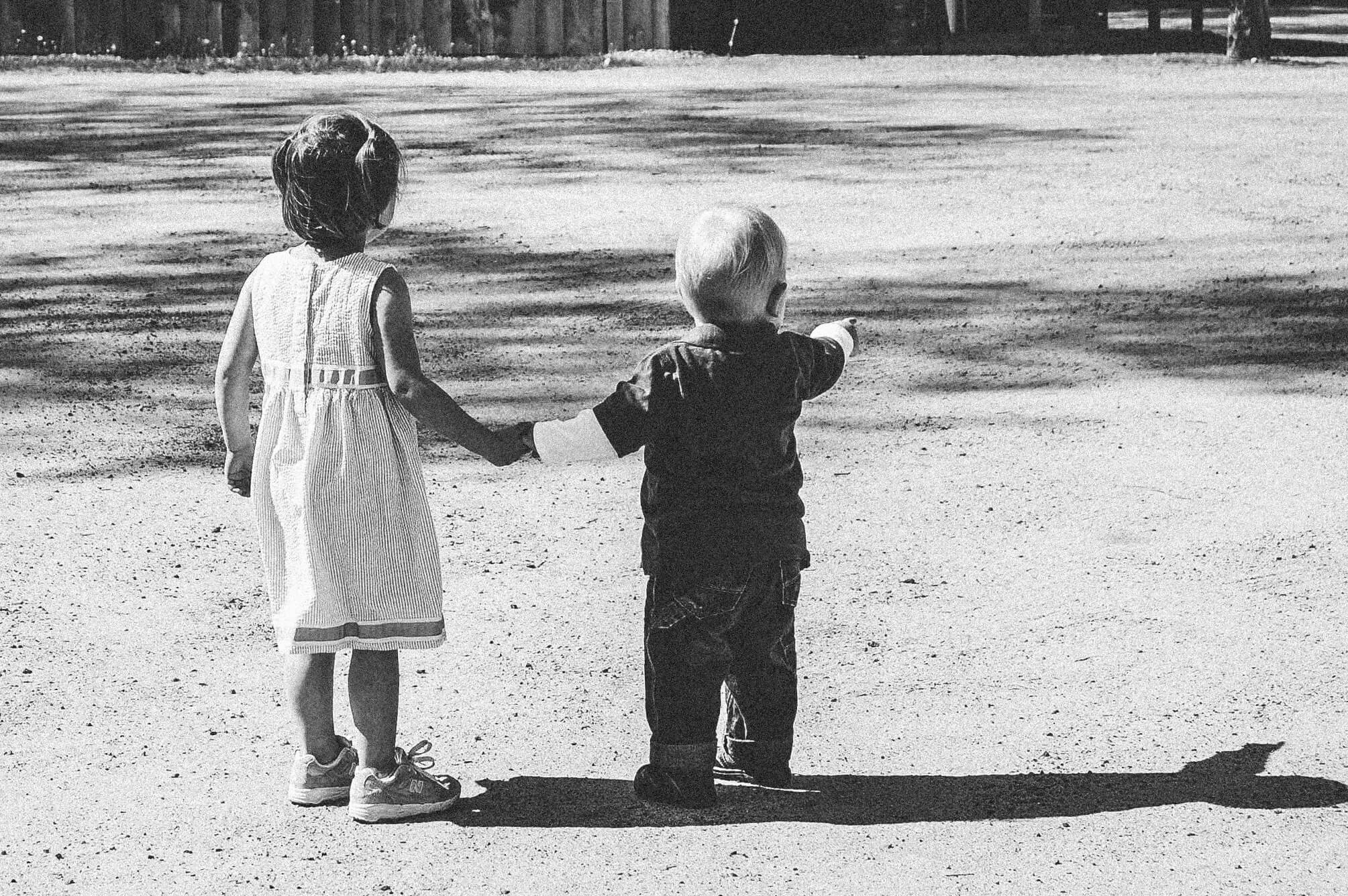 Black and white image of a white boy and girl holding hands. Photo is taken from behind them, you cannot see their faces.