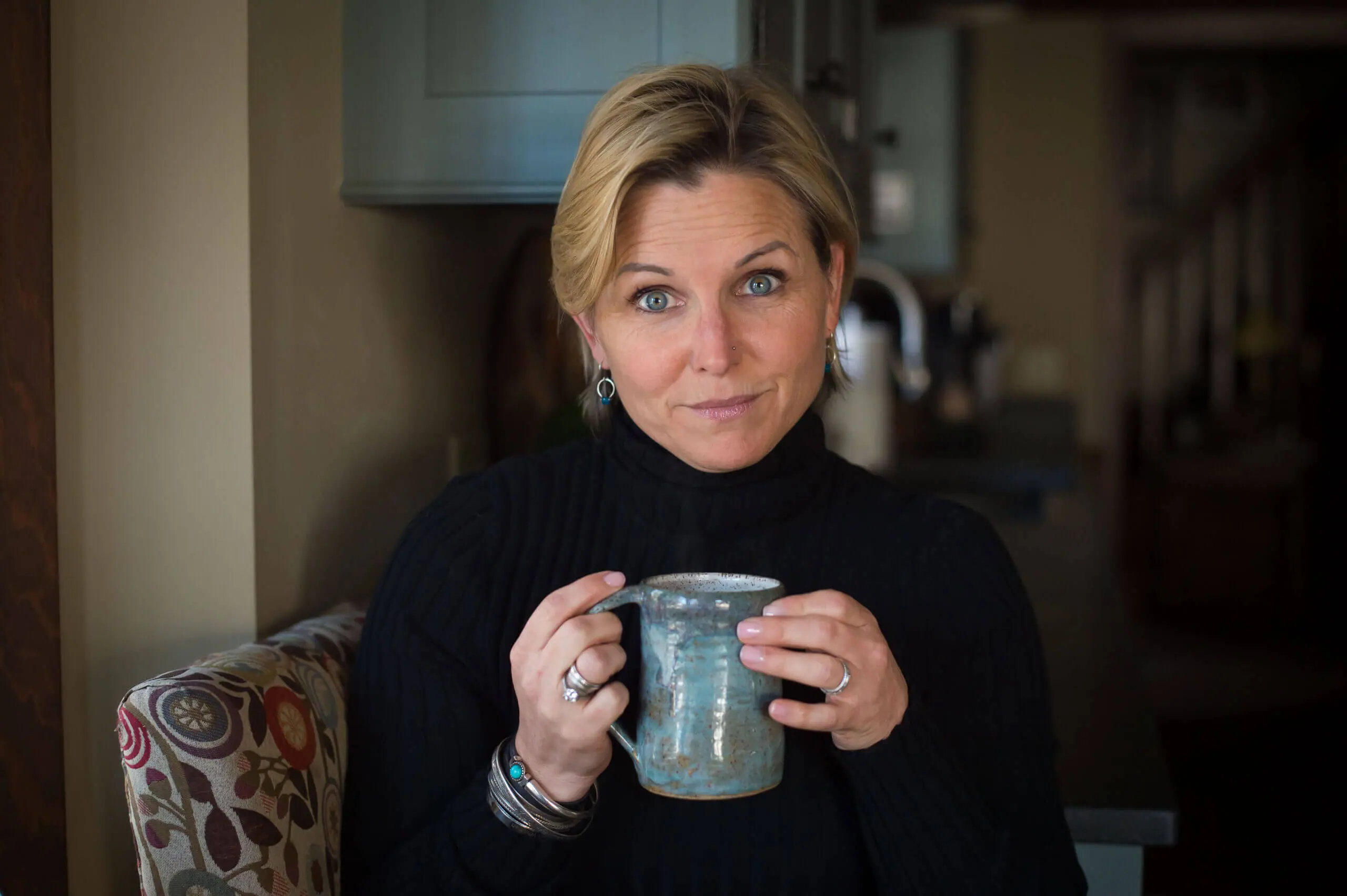 Color photo of a white 40-something year old woman with short blond hair holding a coffee cup with a smirk on her face.