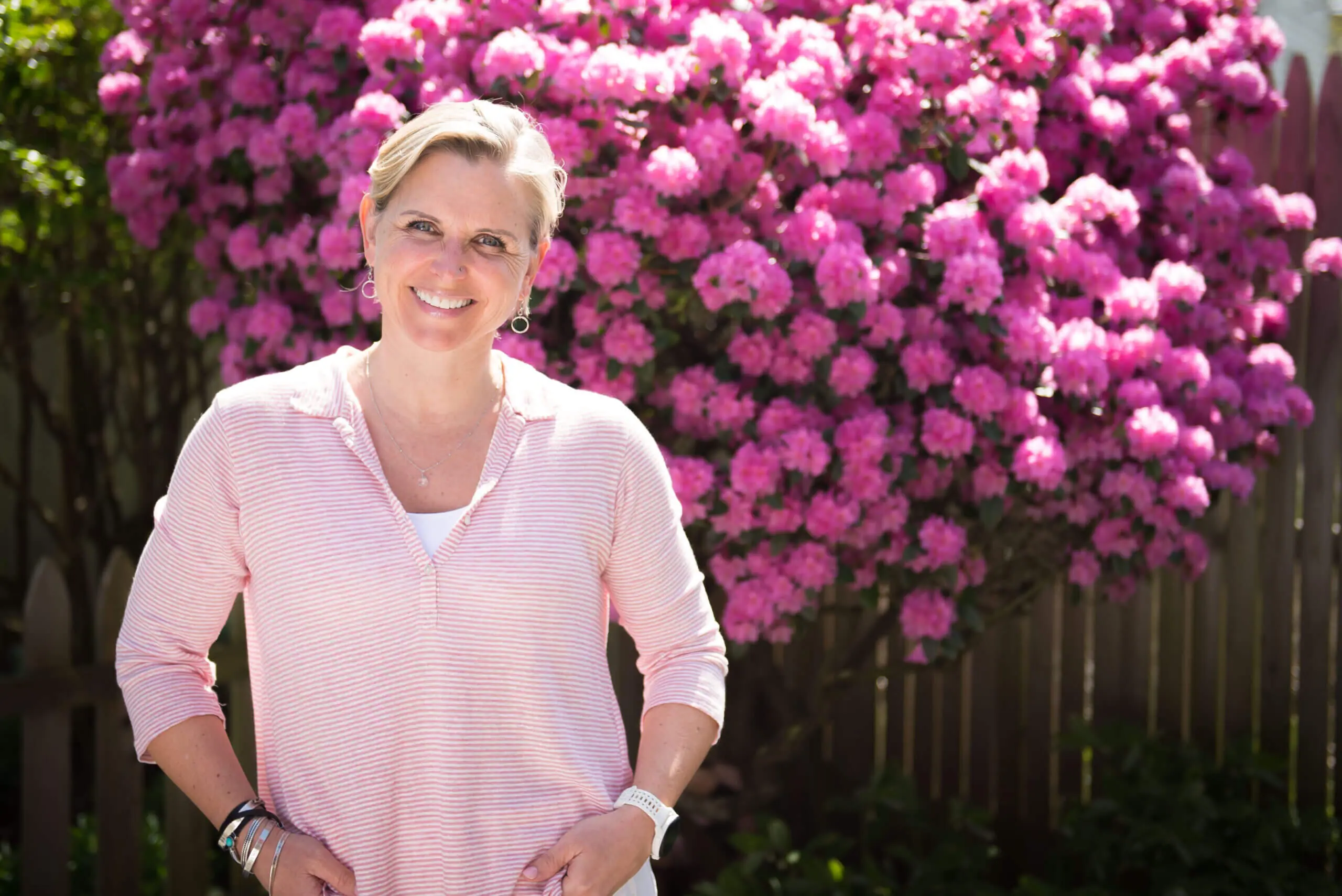 White middle aged woman in a pink shirt in front of a pink flowering bush.