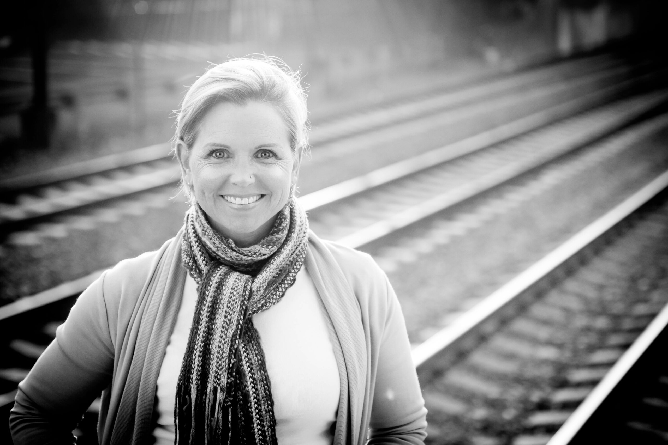 Black and white image of white 50 year old woman with short blond hair smiling with a scarf on and the sun behind her