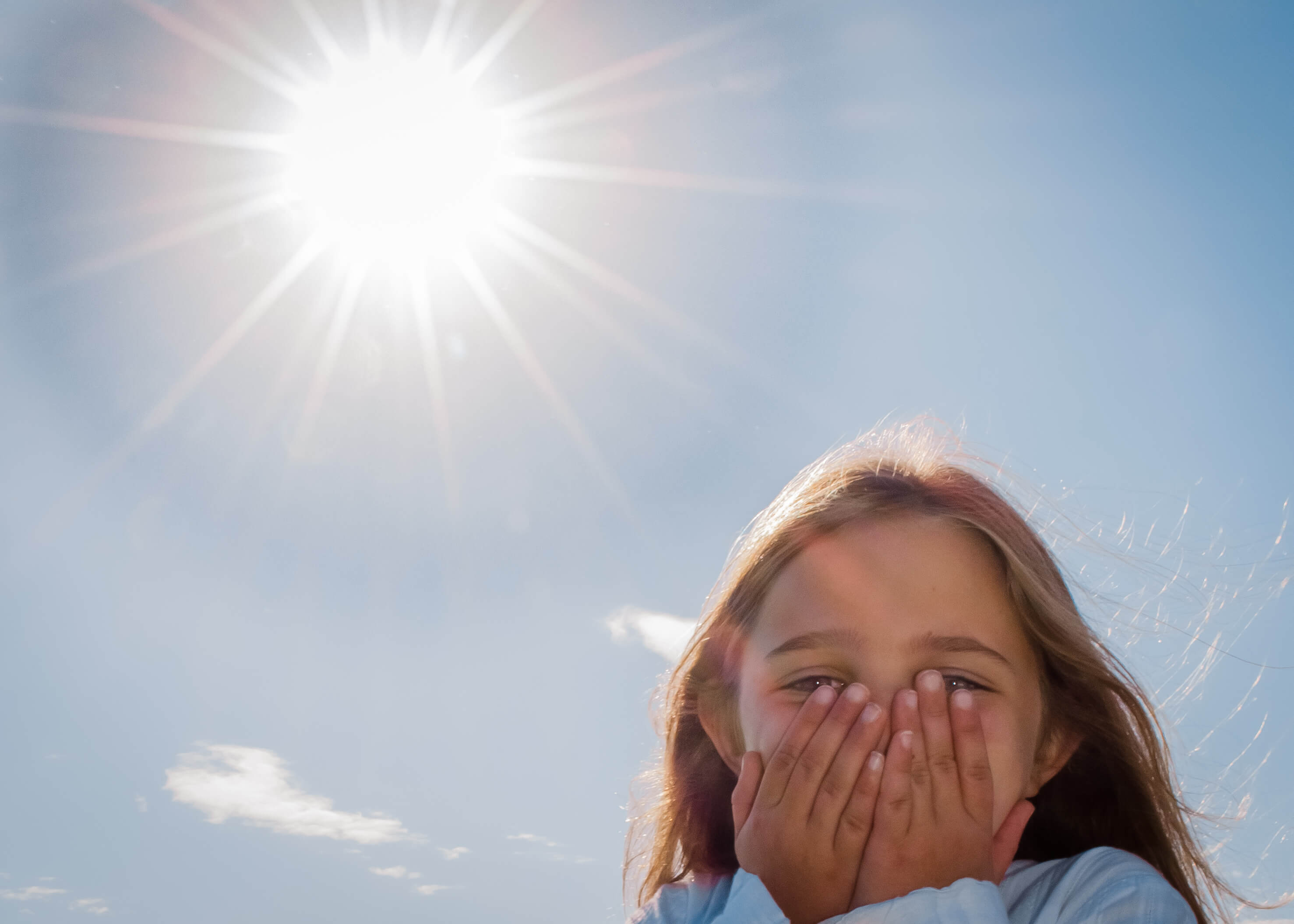 A color photo of a bright sun in the upper left corner and a little white girl with her hands over her mouth, smiling, in the lower right quadrant of the picture.