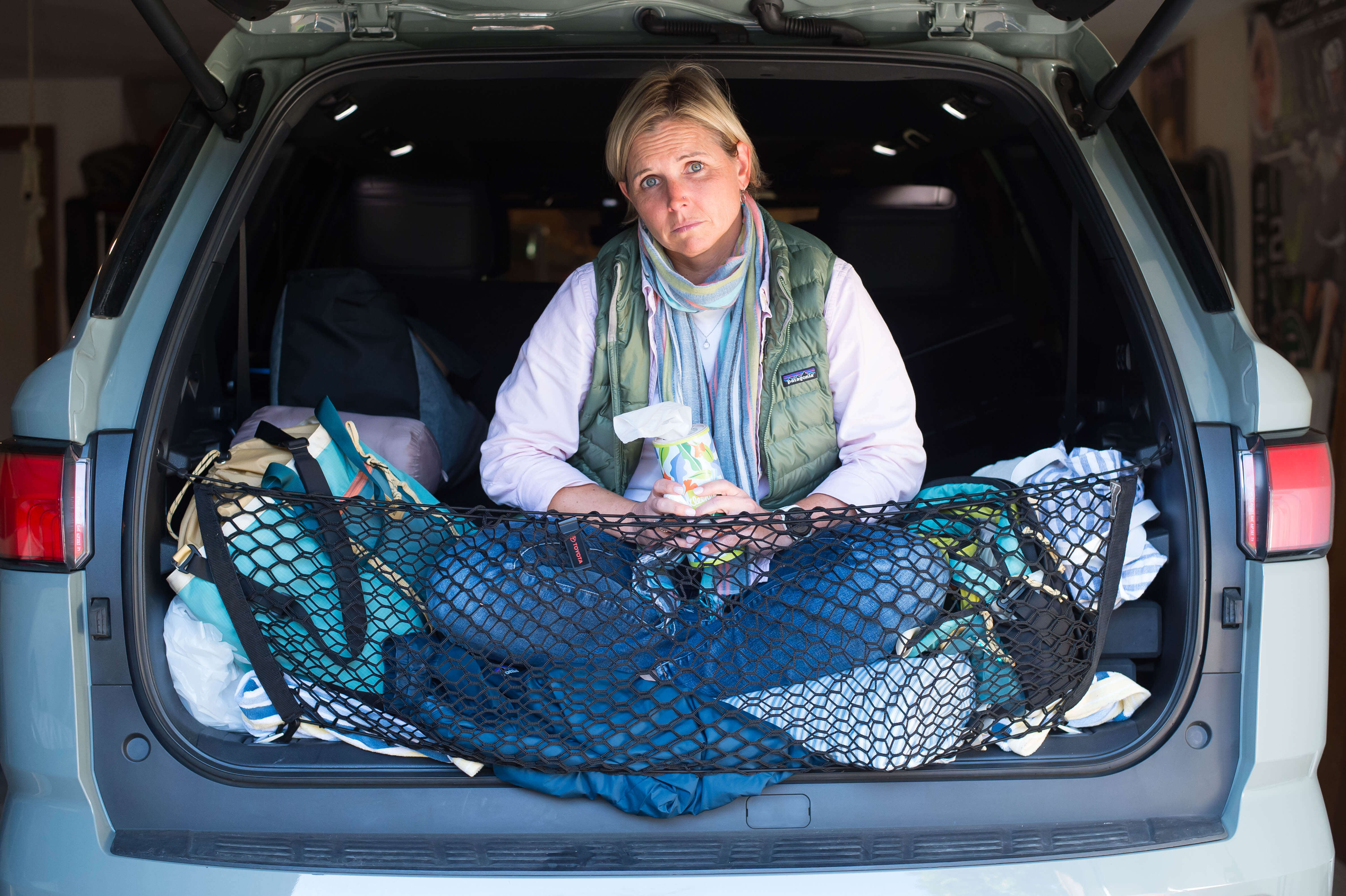 Middle aged white woman in the back of an opened tailgate on an SUV, looking sad with a box of tissues in her hands.