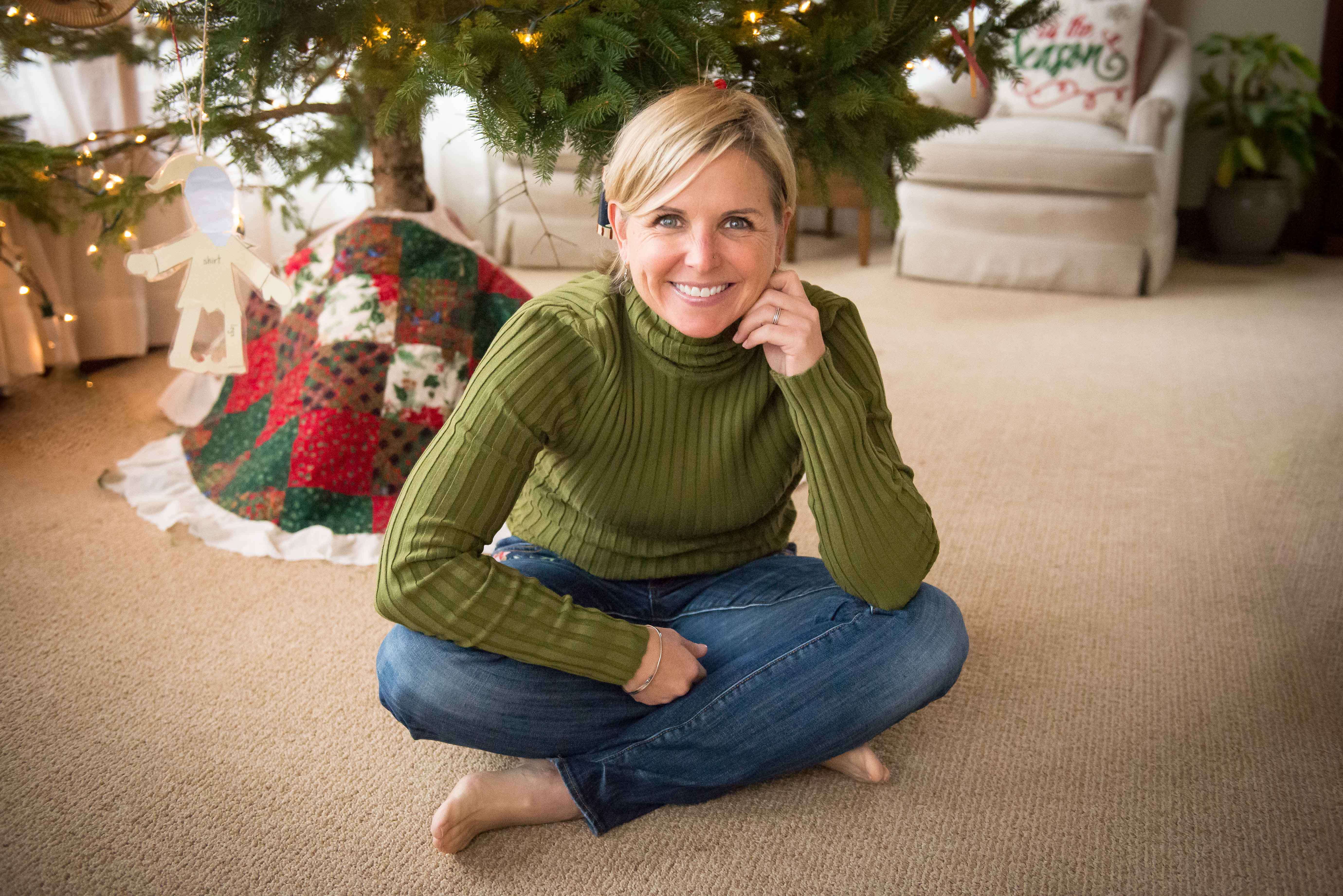 White middle aged women sitting on the floor in a green sweater with a Christmas tree behind her.