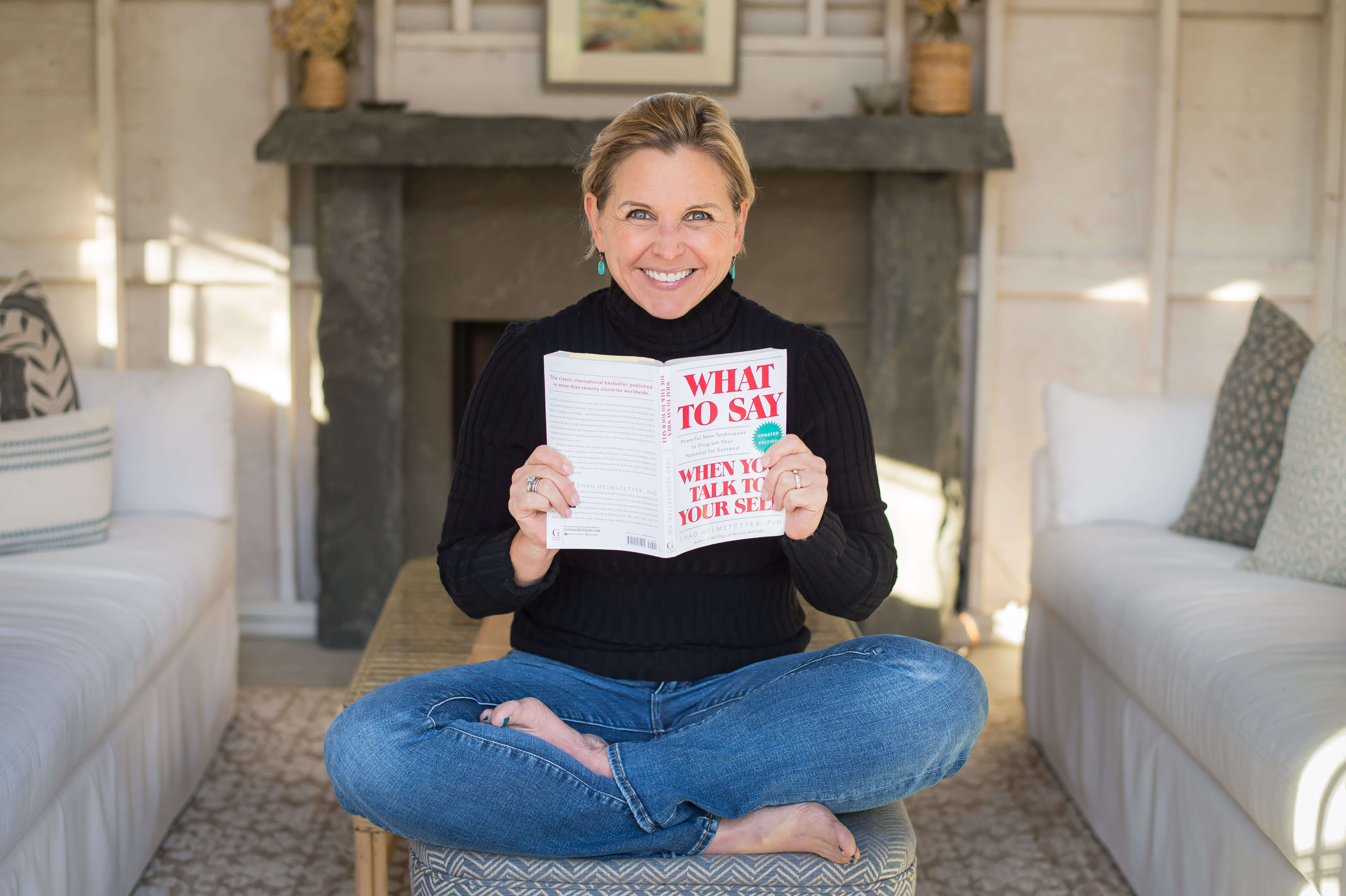 White 50 year old woman sitting crossed legged on a stool holding the book: What To Say When You Talk To Yourself by Shad Helmstetter.