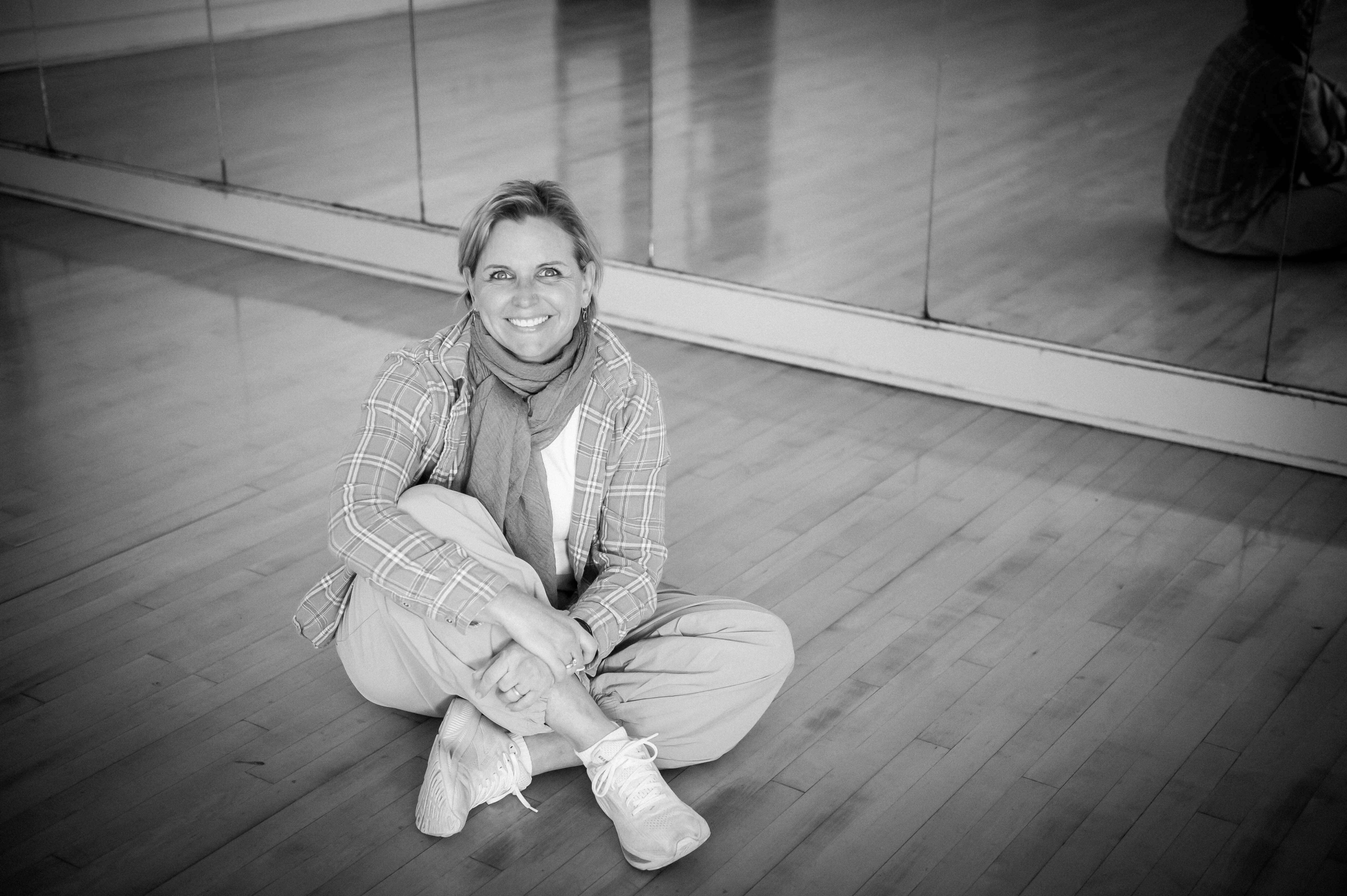Black and white image of a 50 yo white woman sitting on a dance studio floor with a mirror in the background.