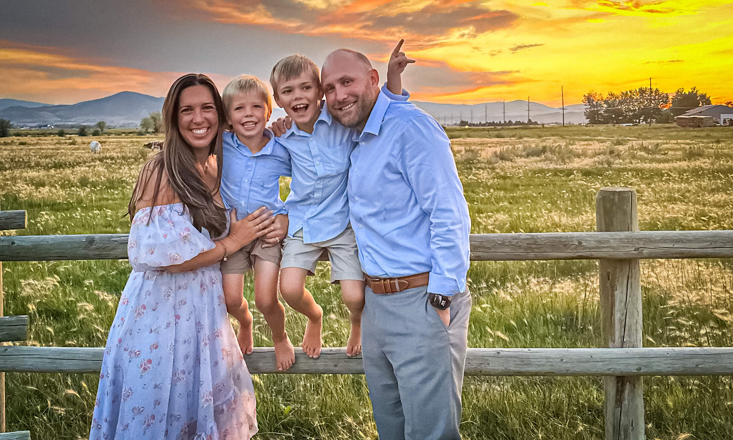 Color photo of a white family sitting on a split-rail fence in front of a Montana sunset: Mom, Dad, and two young boys.