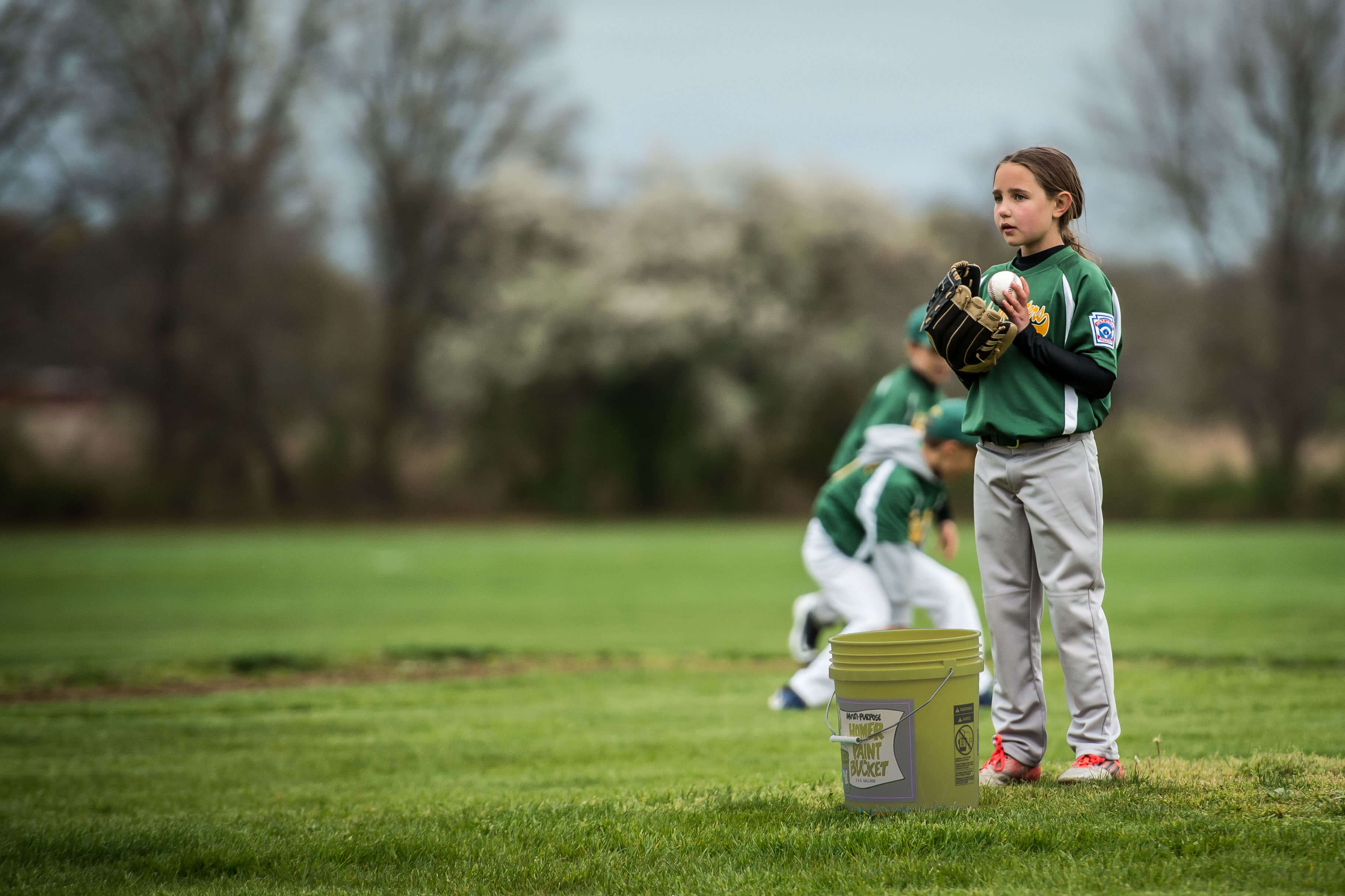 Image of a young white girl in a baseball uniform on a pitching mound with a glove on her right hand and a baseball in her left hand.