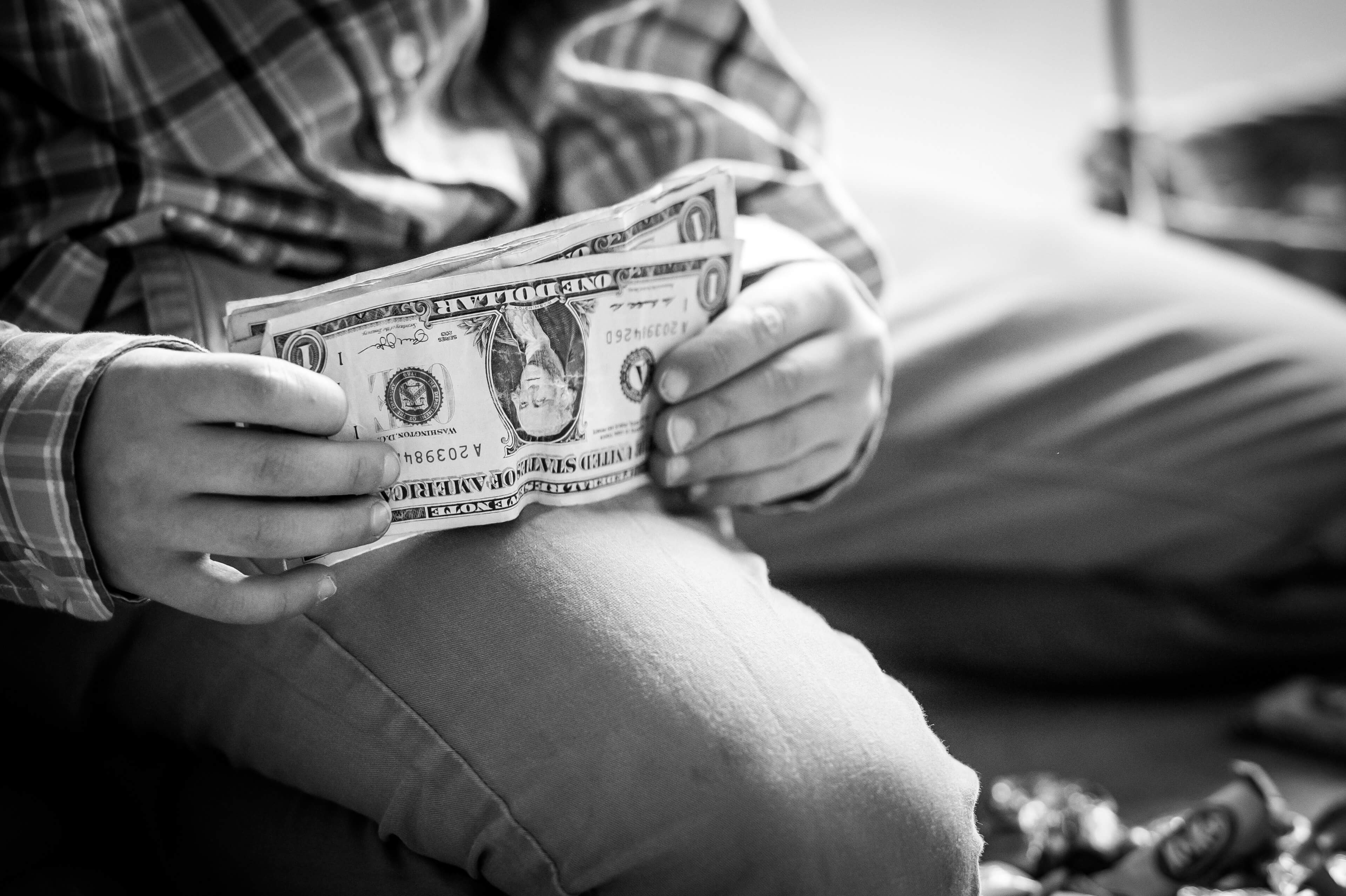 Black and white image cropped in to see a six year old hands straightening out a bunch of one dollar bills on his thigh.