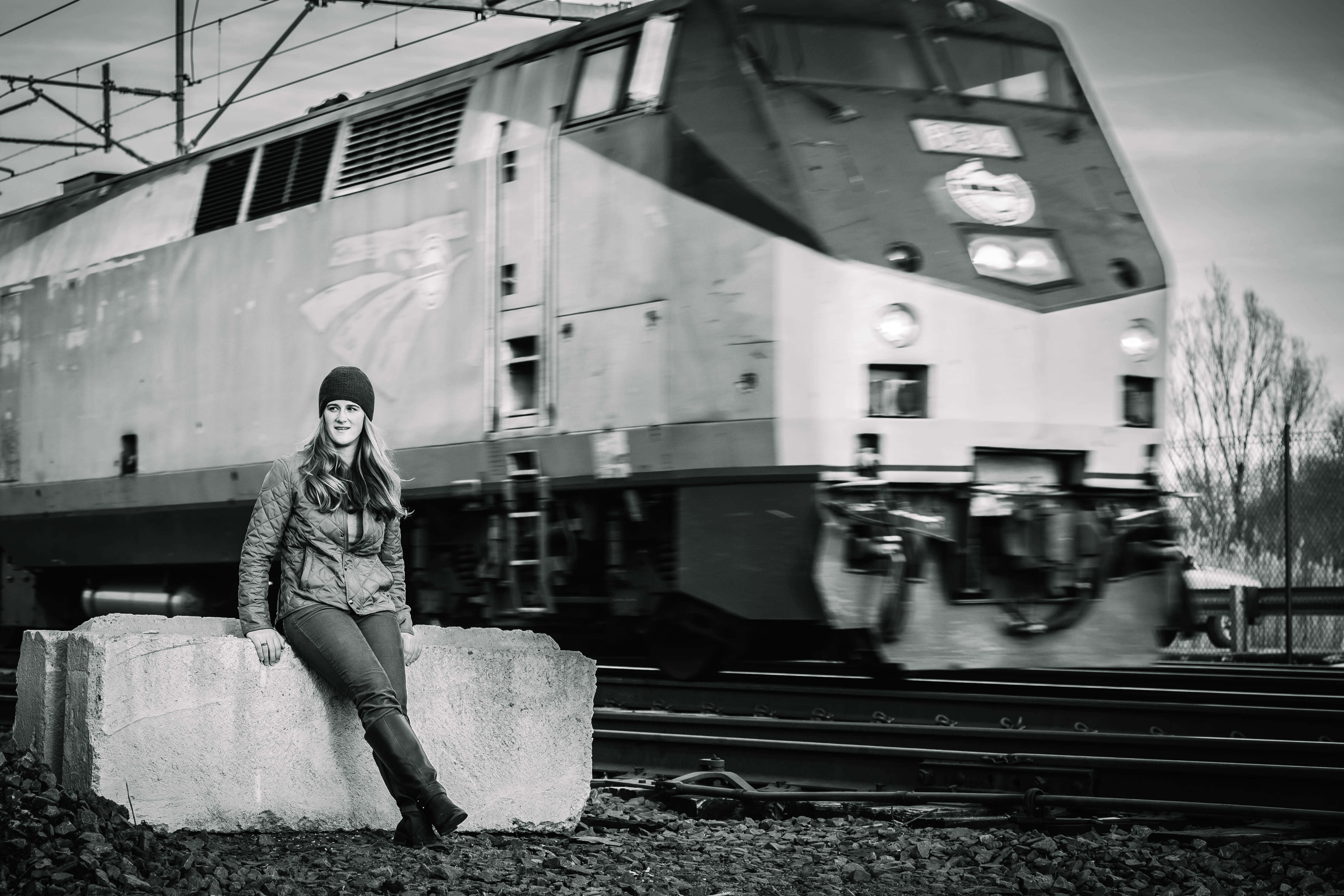 Image of a young white woman calmly leaning on a large concrete barrier as an Acela train speeds by her.