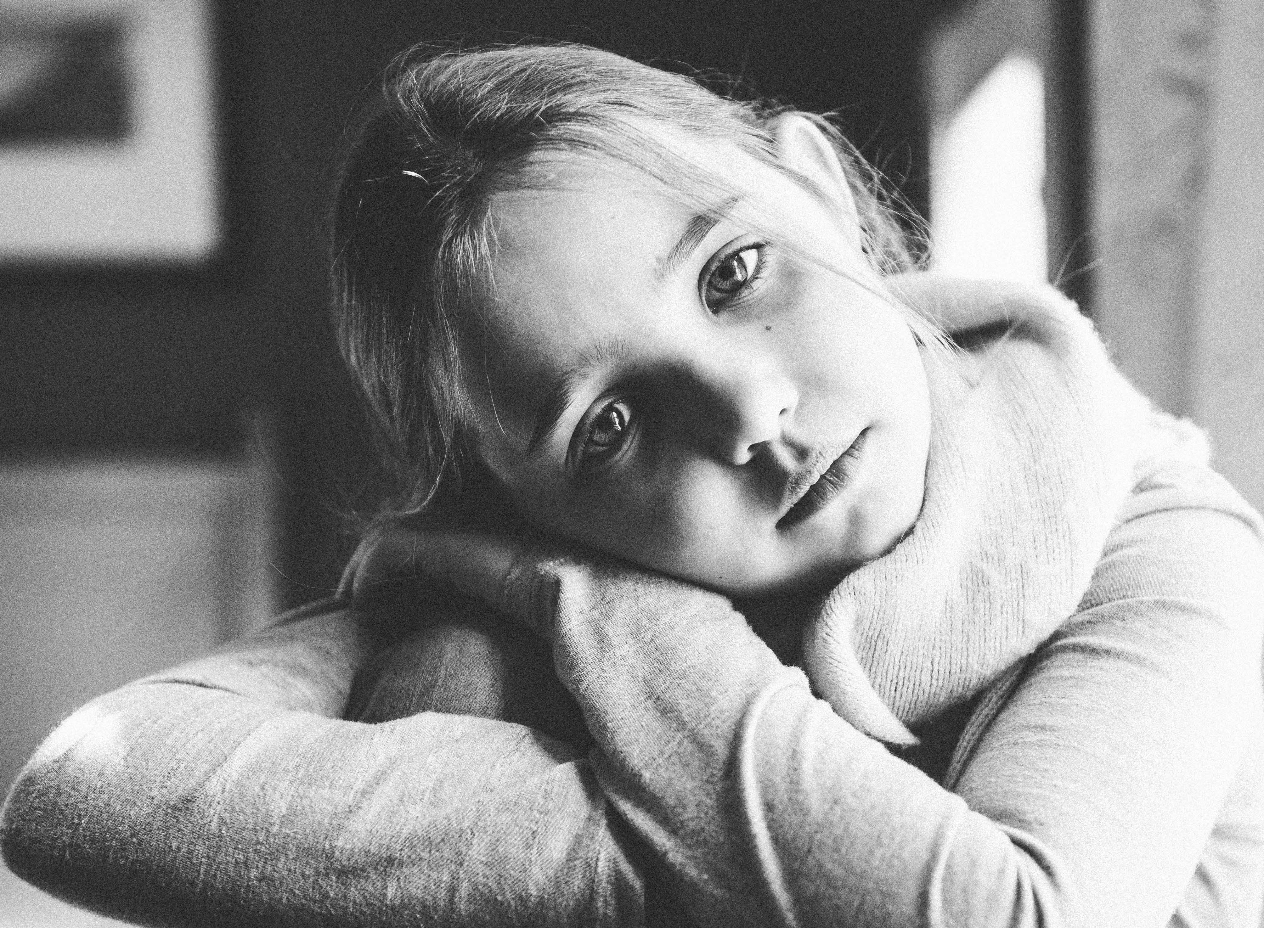 Black-and-white image of a young girl with a reflective expression resting her head on folded arms.