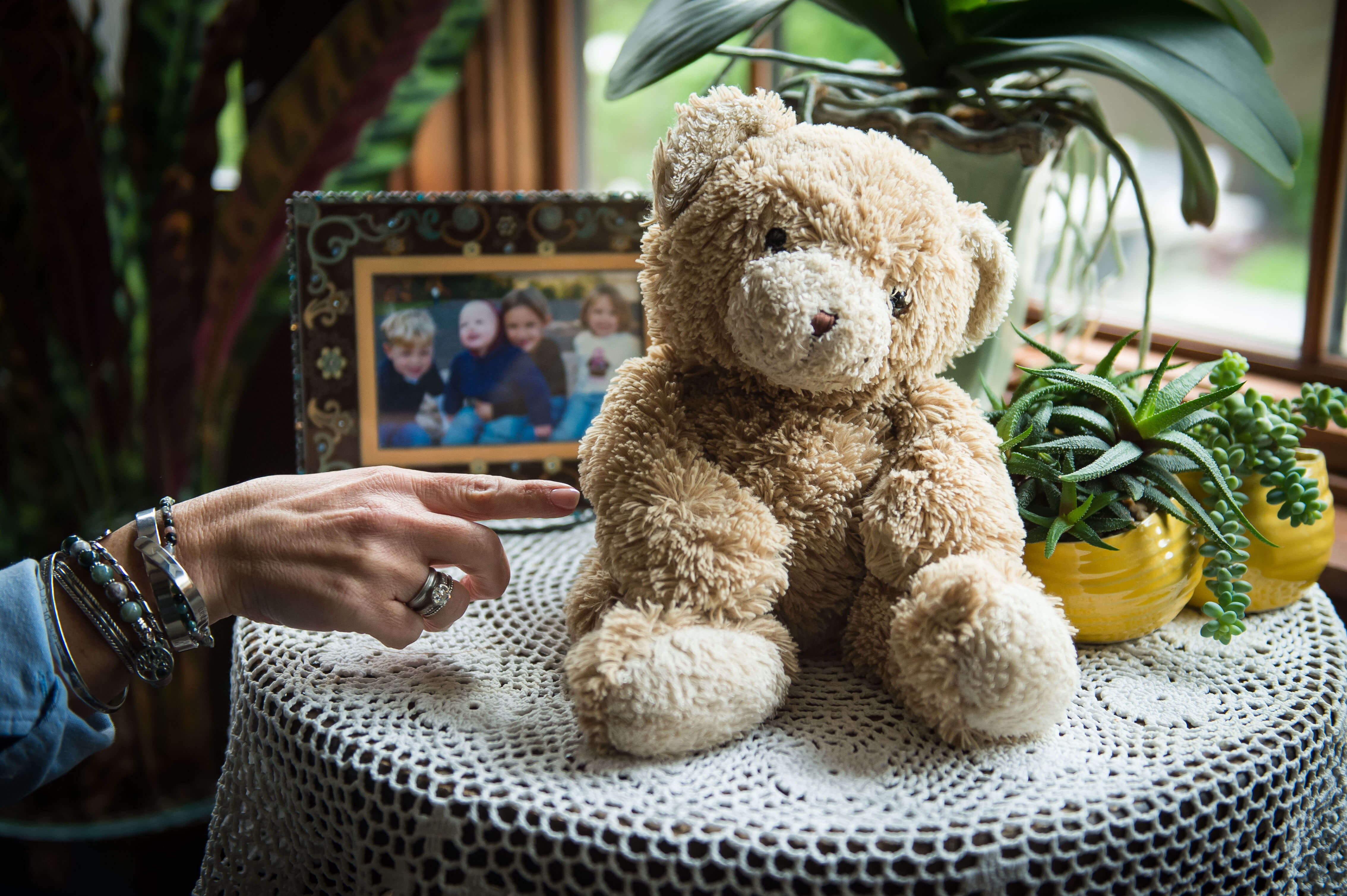 Color photo of a teddy bear on a side table with a white woman's hand about to poke its belly.